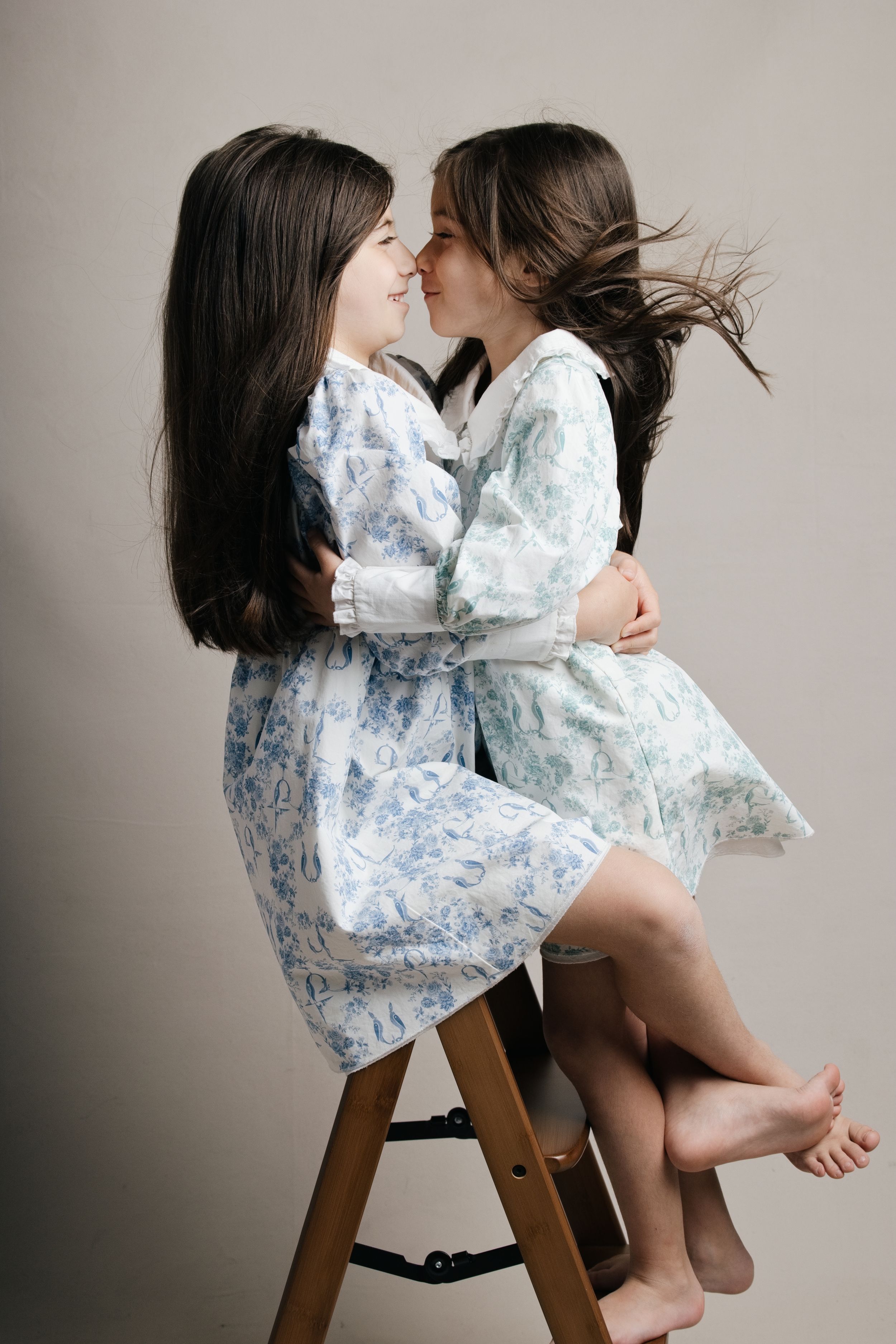 Two young sisters in matching blue floral dresses embracing and smiling at each other on a wooden stool.