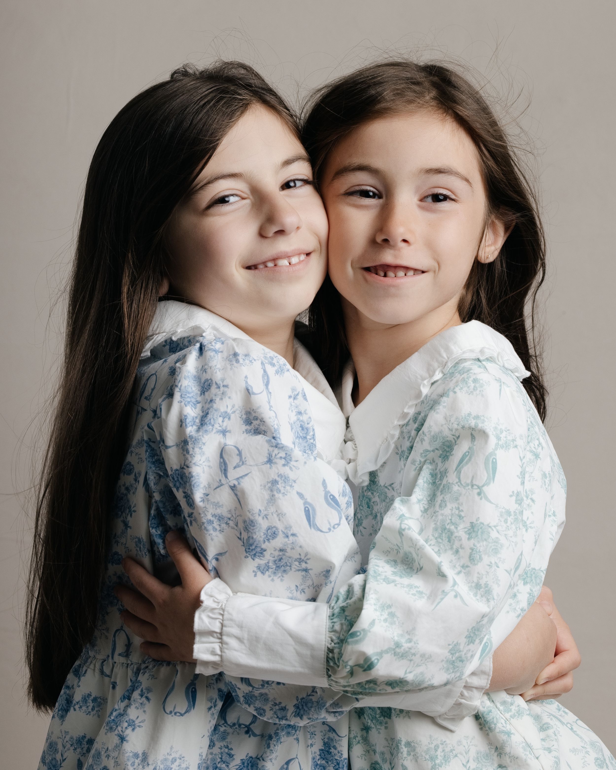 Two young girls in matching floral dresses hugging and smiling in a studio portrait photo.