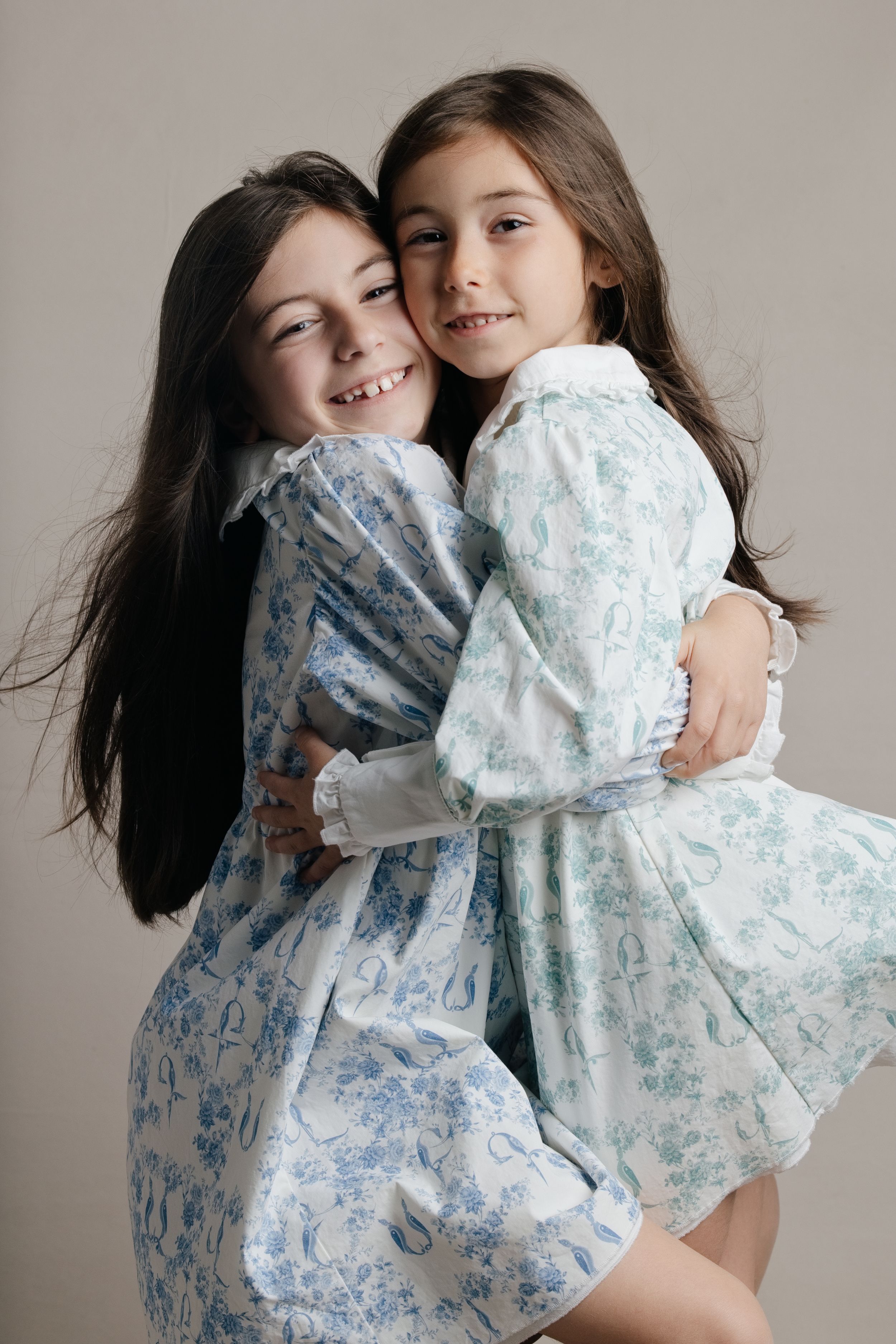 Two young girls in matching floral dresses hugging and smiling in a studio photo shoot.