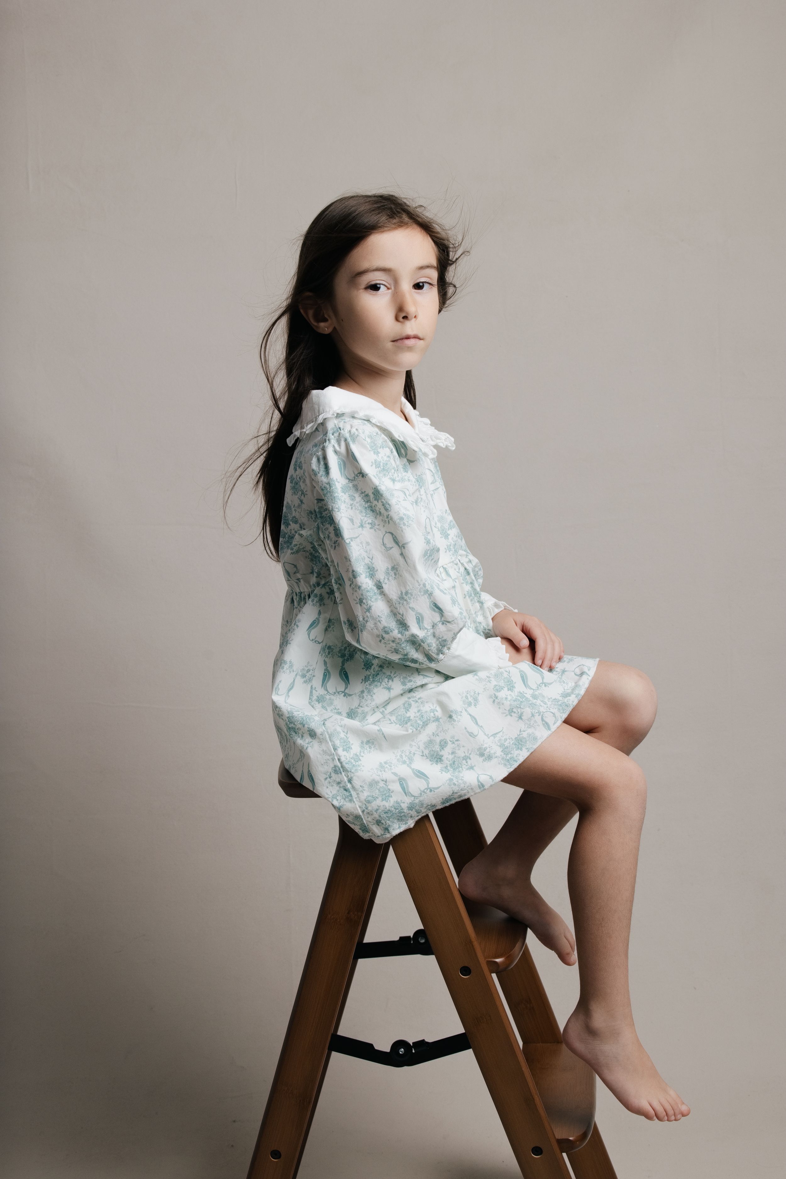 Young girl in a light blue floral dress with white collar sits barefoot on a wooden stool in a studio.