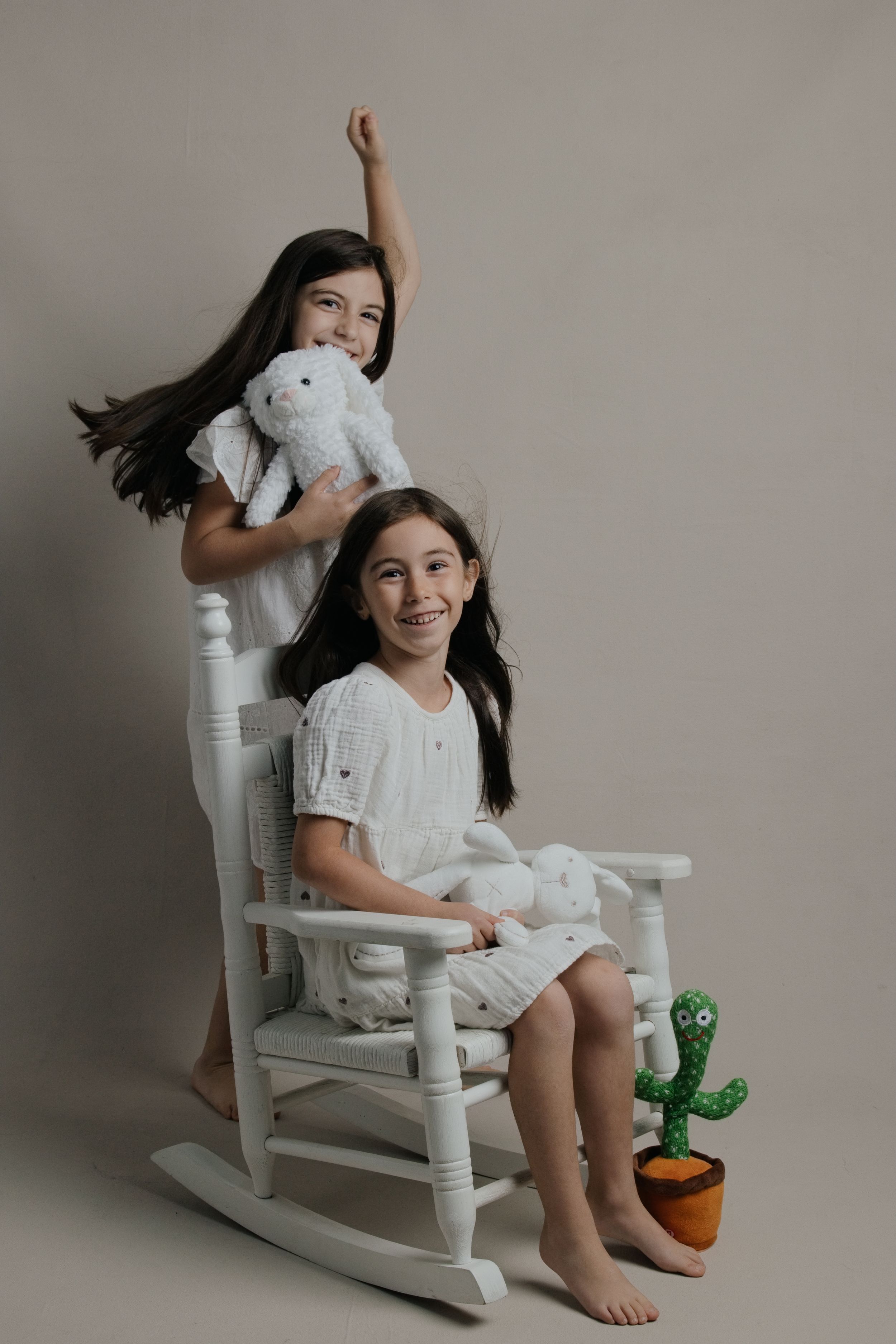 Two young girls in white outfits pose with stuffed animals and a cactus toy on a white rocking chair.