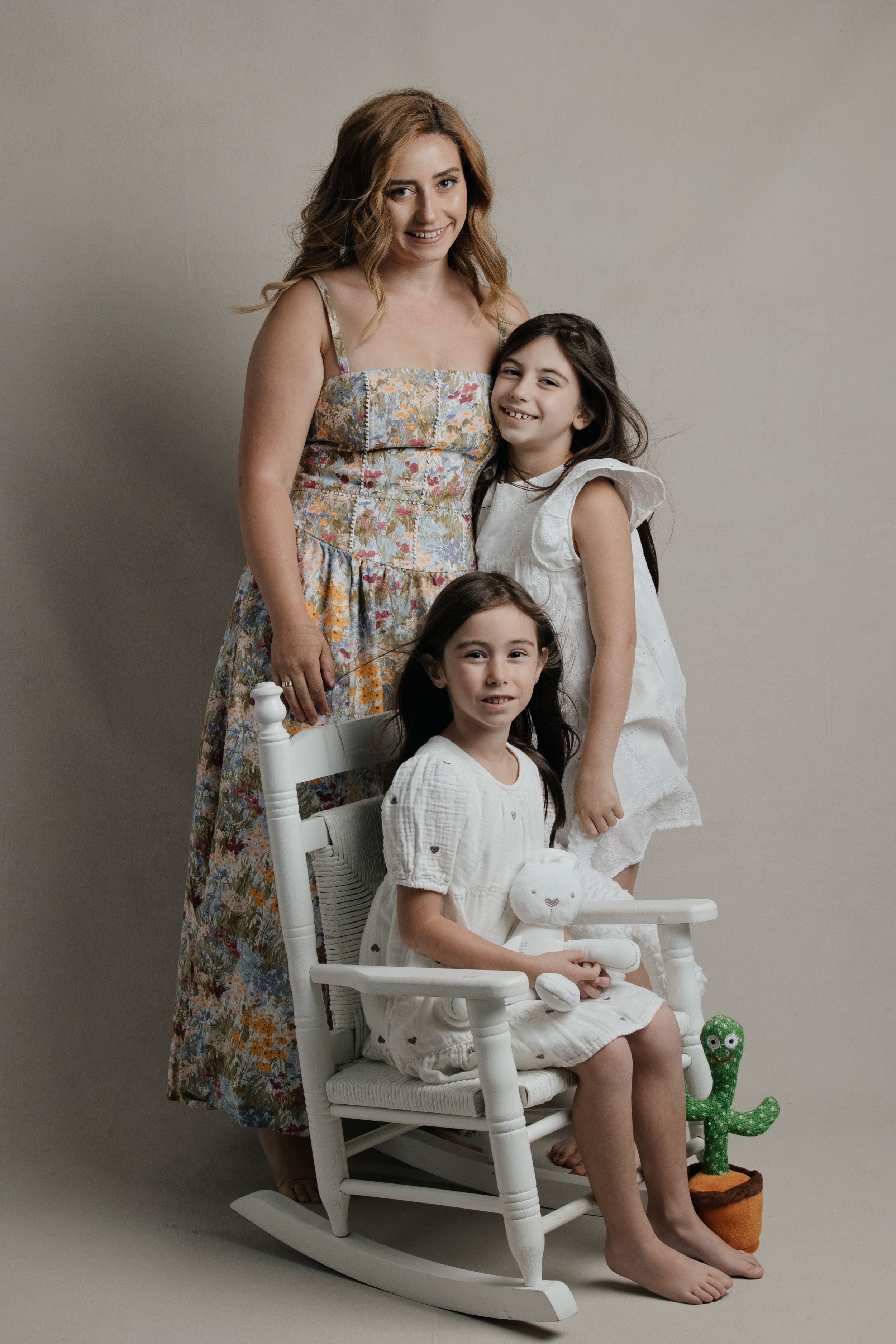 Woman in floral dress posing with two young girls in white outfits in studio setting.