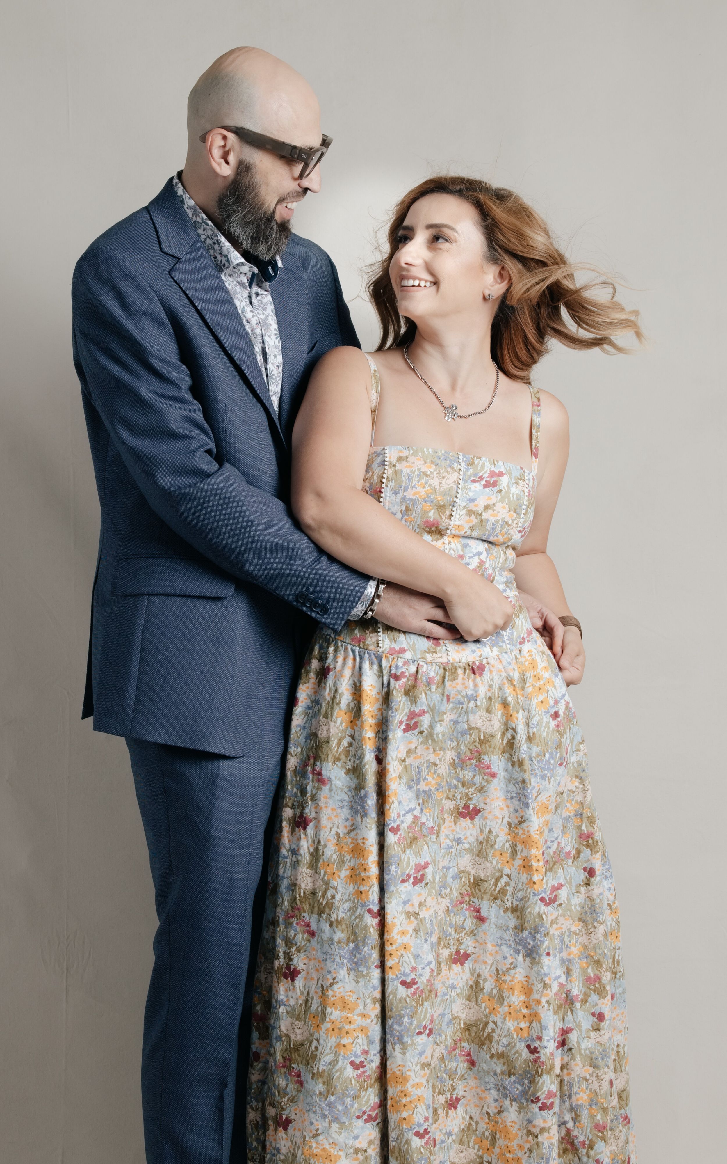 A bald bearded man in a blue suit embracing a smiling woman in a floral maxi dress against a gray backdrop.