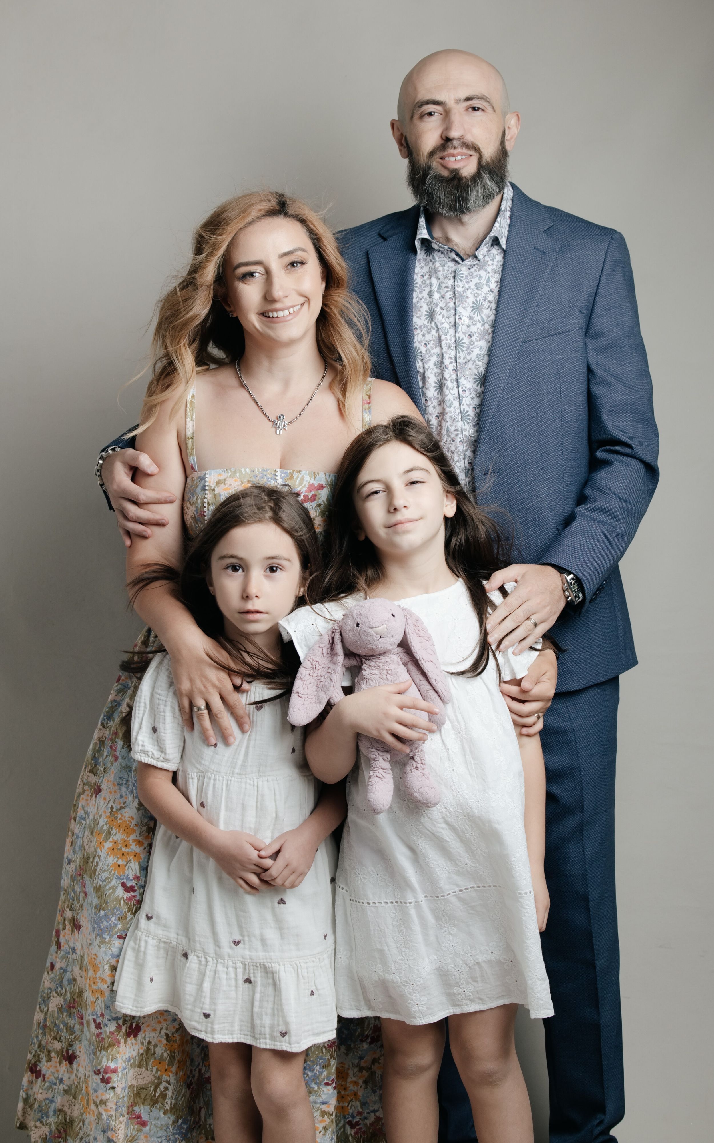 A family of four poses together, parents with two young girls in white dresses.