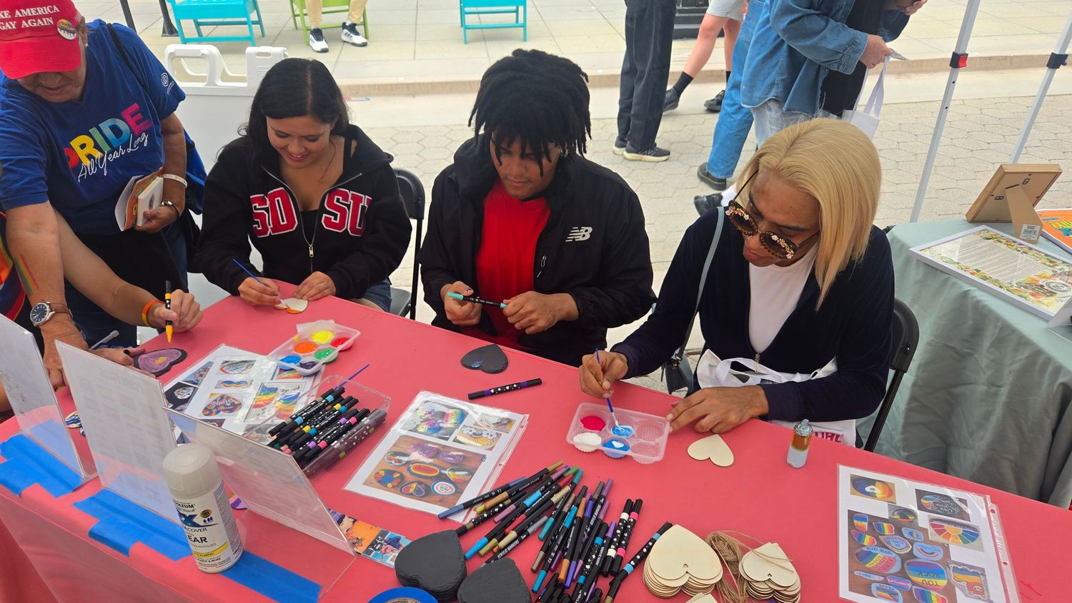 People signing autographs at a colorful event table with markers and photos displayed on a red tablecloth.