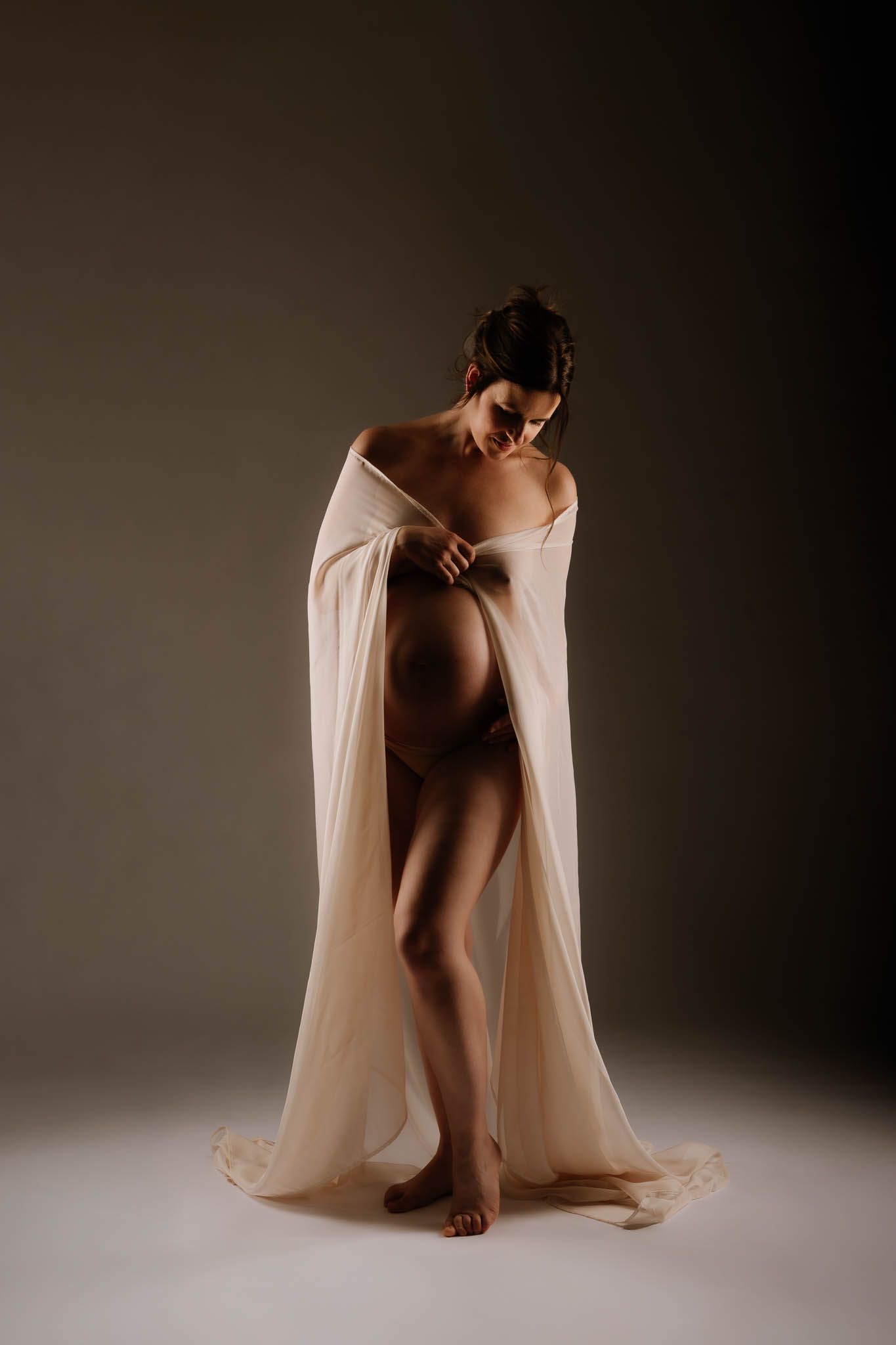 Elegant pregnant woman draped in sheer ivory fabric poses gracefully in a softly lit studio maternity portrait.