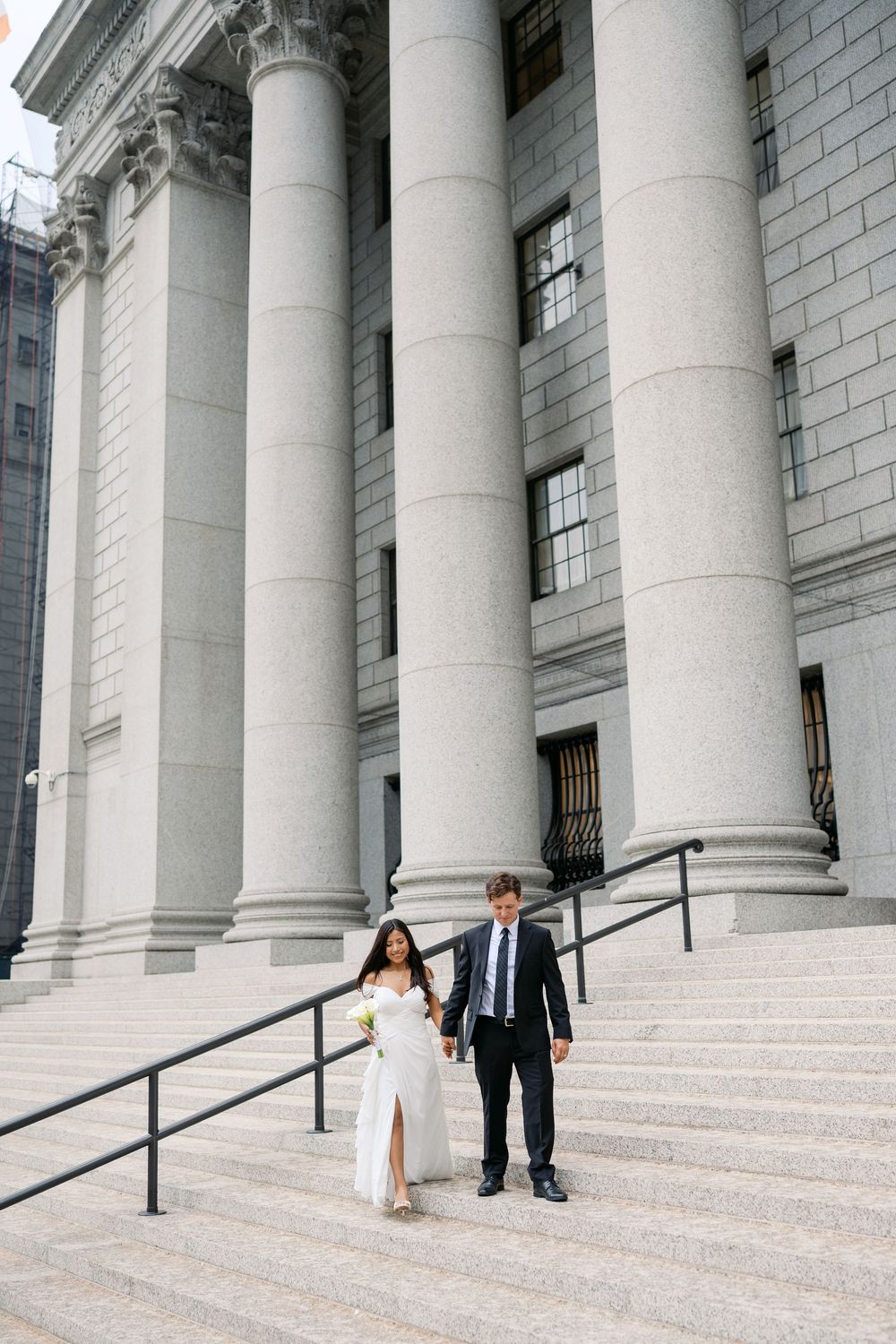 Bride and groom walk down steps of grand courthouse with large stone columns on their wedding day.