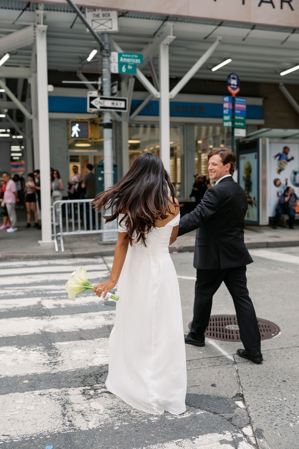 Bride in white gown holding bouquet crosses NYC street with groom in black suit near busy urban intersection.