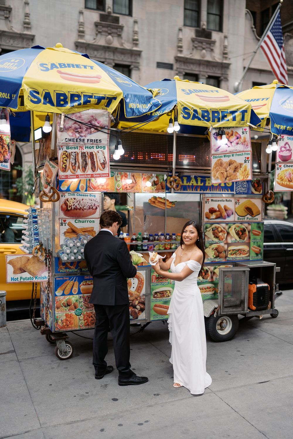 Bride and groom stopping at a Sabrett hot dog cart on a New York City street in wedding attire.