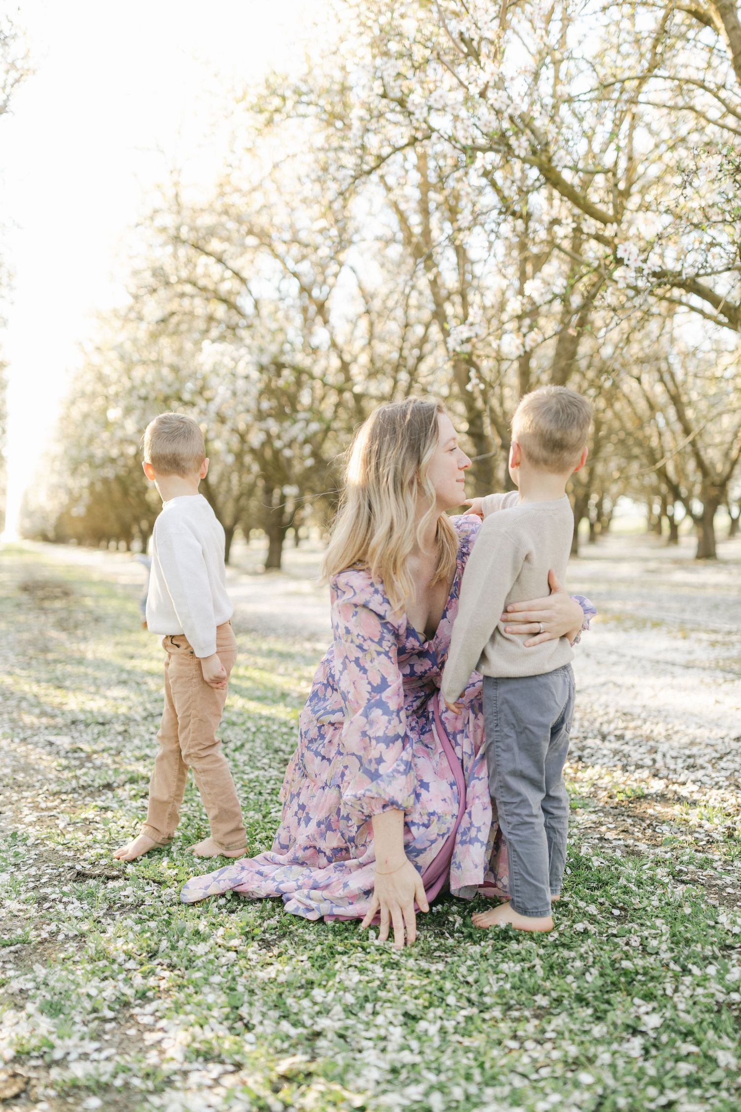 A mother in a floral dress sits on grass hugging her two young sons in a sunlit orchard with blooming trees, comforting them