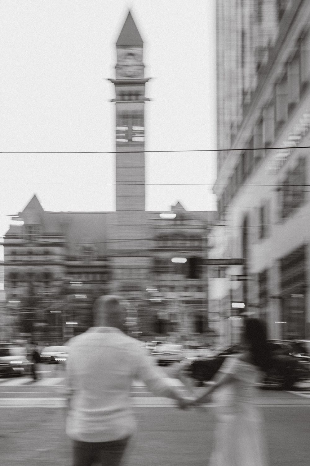 Couple walking hand in hand past a clock tower in downtown Toronto, motion-blurred black and white.
