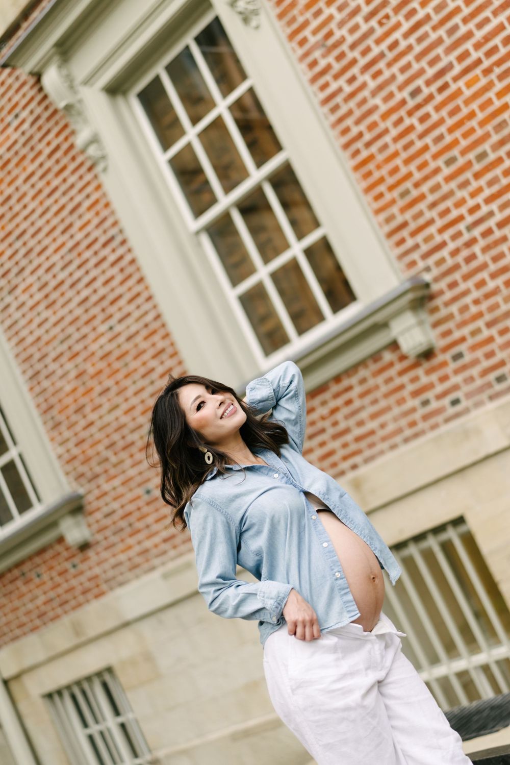 Expectant mother in a denim shirt against a red-brick wall.