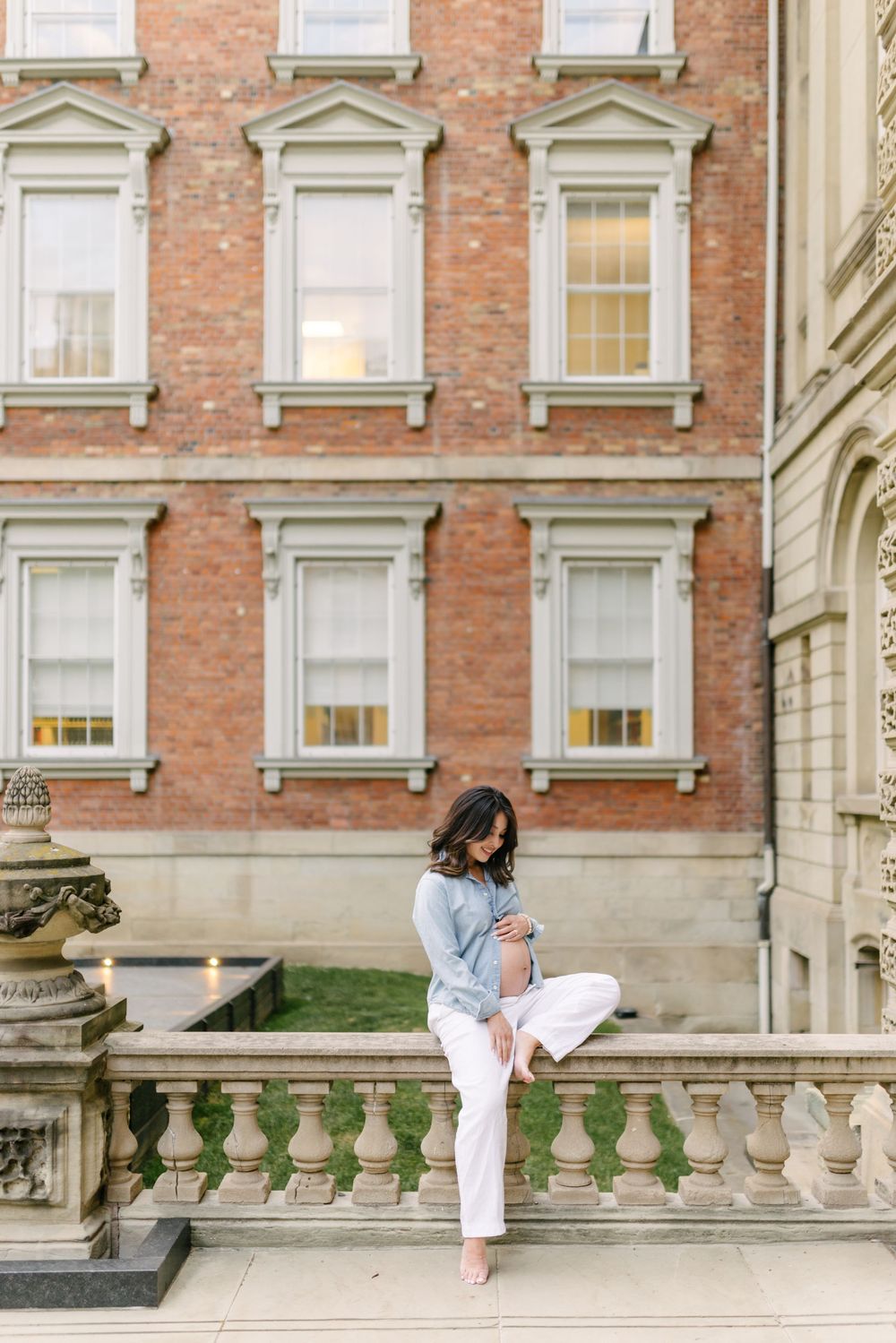 Expectant couple embracing along a balustrade by a red-brick heritage building.