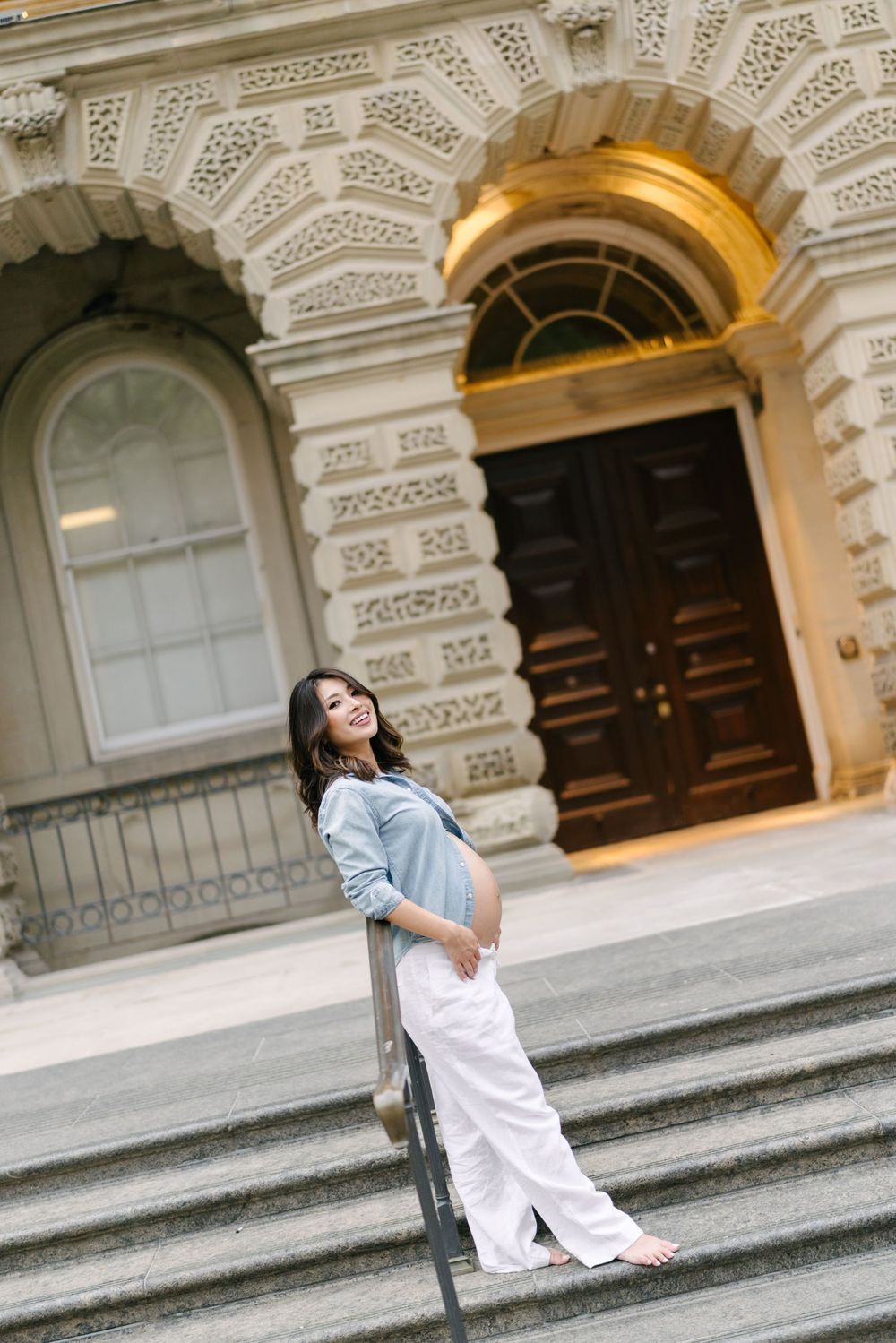 Expectant mother in a denim shirt seated on the steps of a stone building.