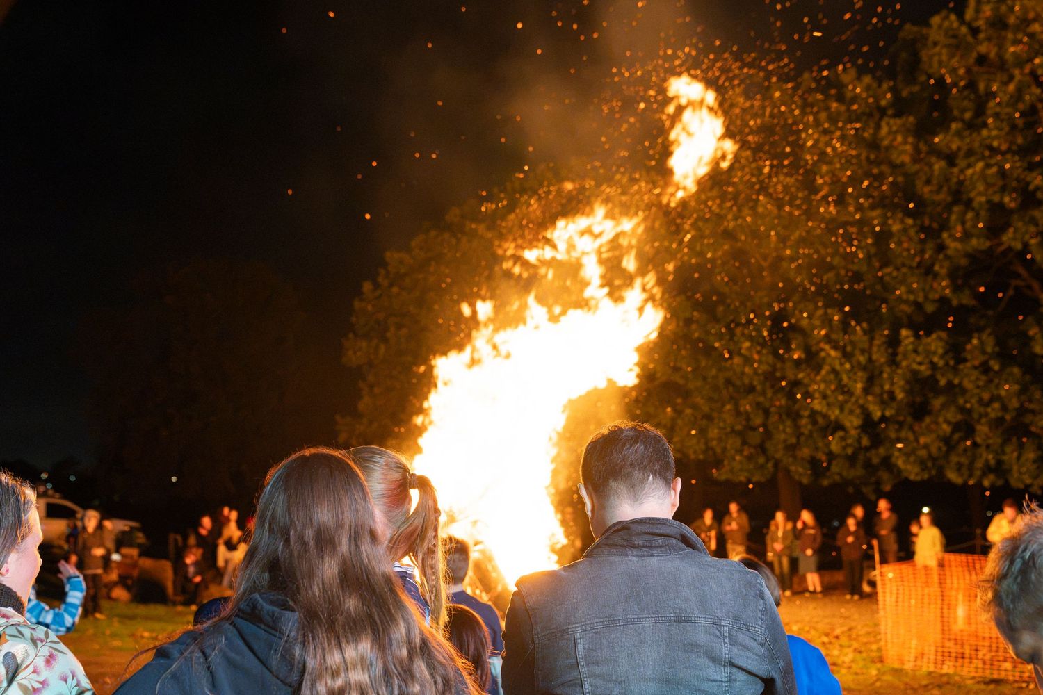 Crowd of people gathered around a large bonfire at night, with flames and sparks illuminating the dark sky.