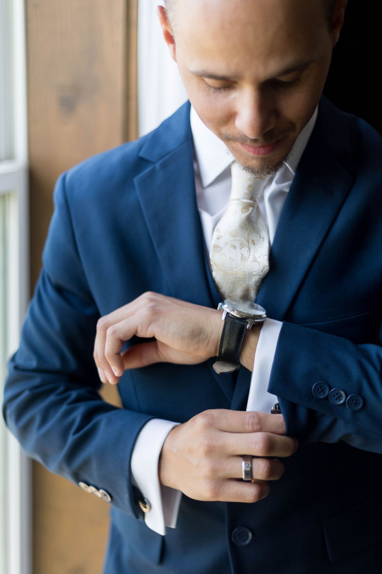 Groom in blue suit adjusting watch with white paisley tie and wedding ring before ceremony.
