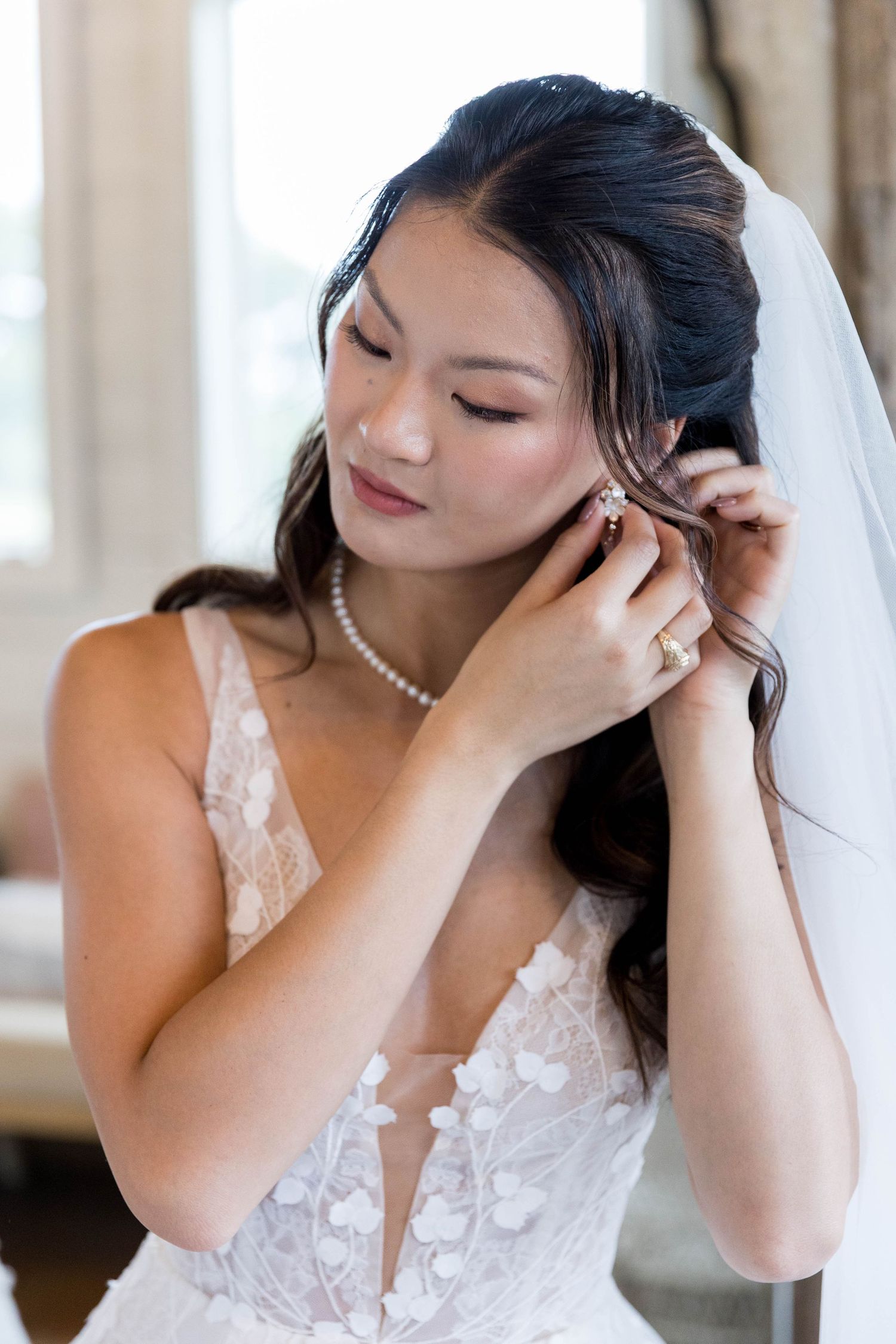 Bride in lace wedding gown adjusting earring, wearing pearl necklace and veil, soft natural light background.