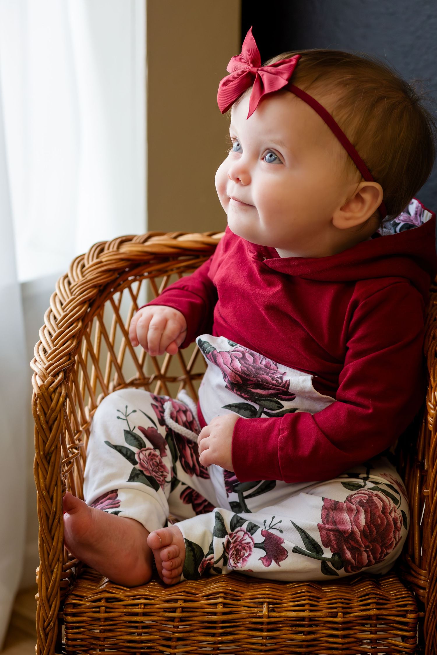 Adorable baby girl in red floral outfit and bow headband sitting in wicker chair, looking up with blue eyes.