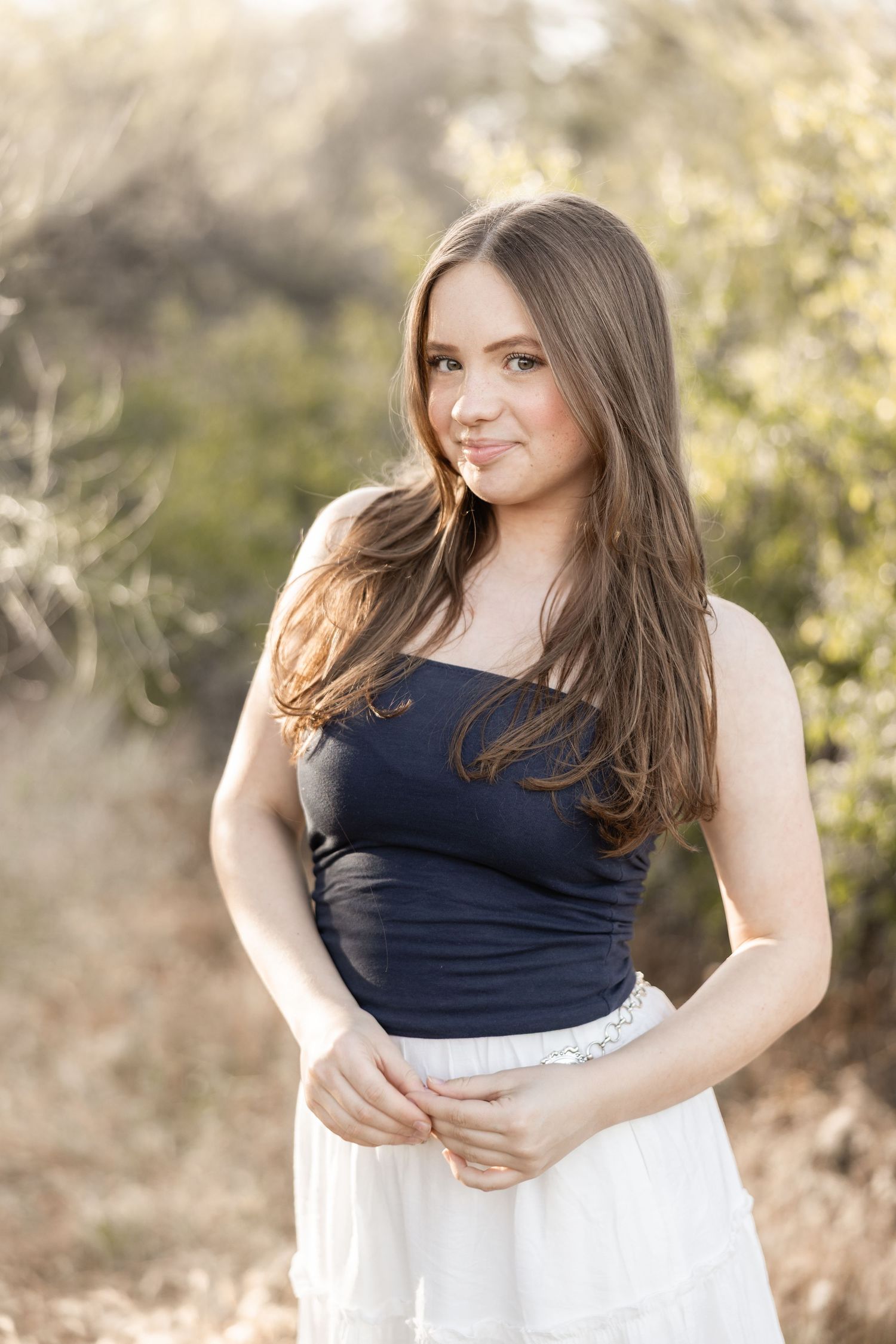 Young woman with long brown hair wearing a navy top and white skirt, smiling outdoors in natural setting.
