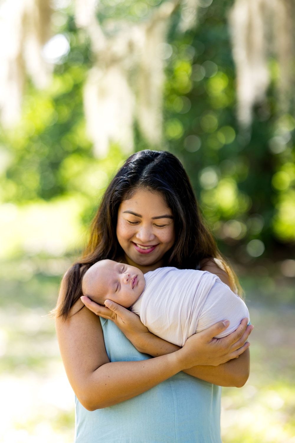 Smiling mother tenderly holds her sleeping swaddled newborn baby outdoors in a lush green park setting.