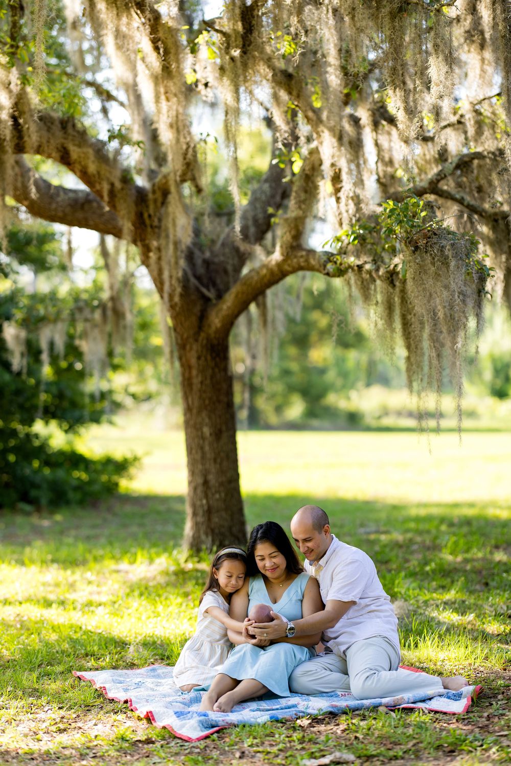Family sitting on blanket under Spanish moss oak tree in sunny park, parents and child enjoying outdoor picnic together.