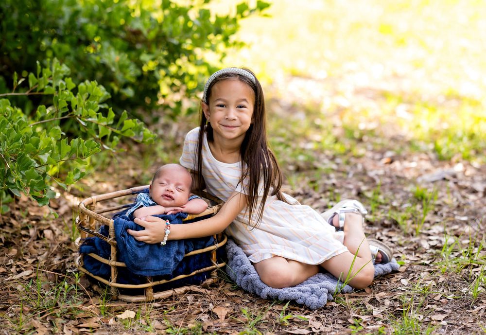 Young girl in white dress sits outdoors cradling a sleeping newborn baby in a small wicker basket.
