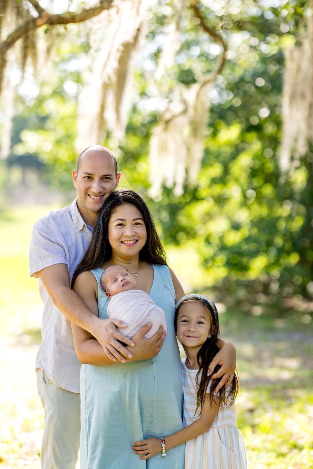 Family of four posing outdoors under Spanish moss trees, parents holding newborn baby and young daughter smiling.