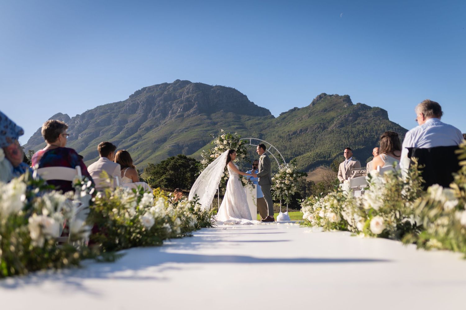 Outdoor wedding ceremony with floral arch, white aisle, guests seated, and stunning mountain backdrop under clear blue sky.