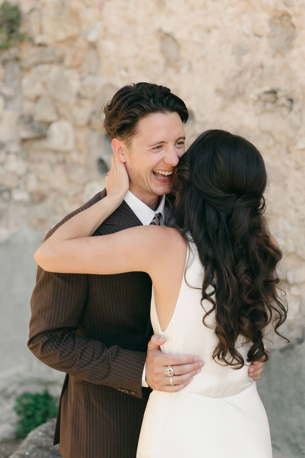 Joyful groom in brown suit laughing as bride in white dress embraces him outdoors near stone wall.