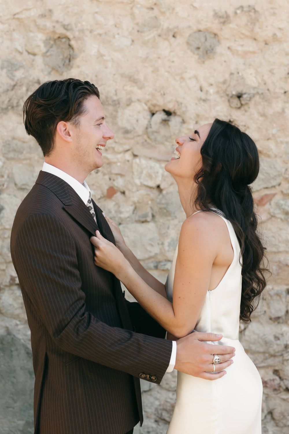 Smiling bride in ivory dress and groom in brown pinstripe suit share a joyful moment against a stone wall.