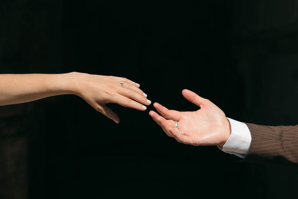 Two hands reaching toward each other against dark background, woman wearing engagement ring, man in suit sleeve.