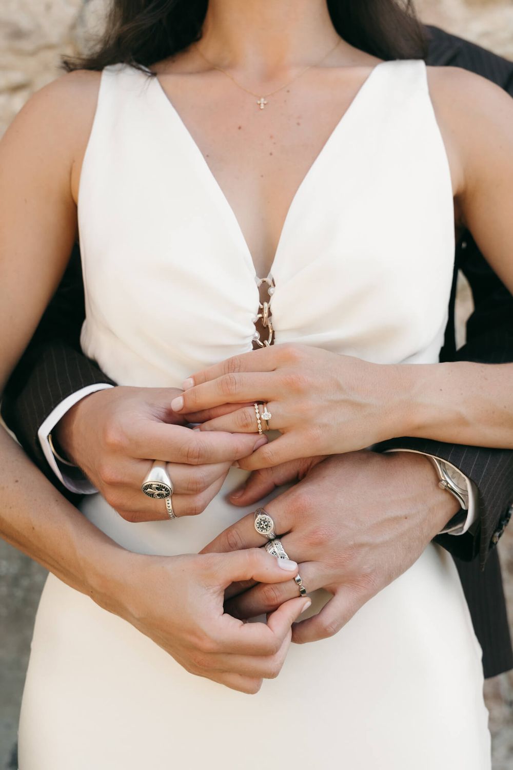 Close-up of a bride and groom holding hands, showcasing elegant rings and a delicate cross necklace.