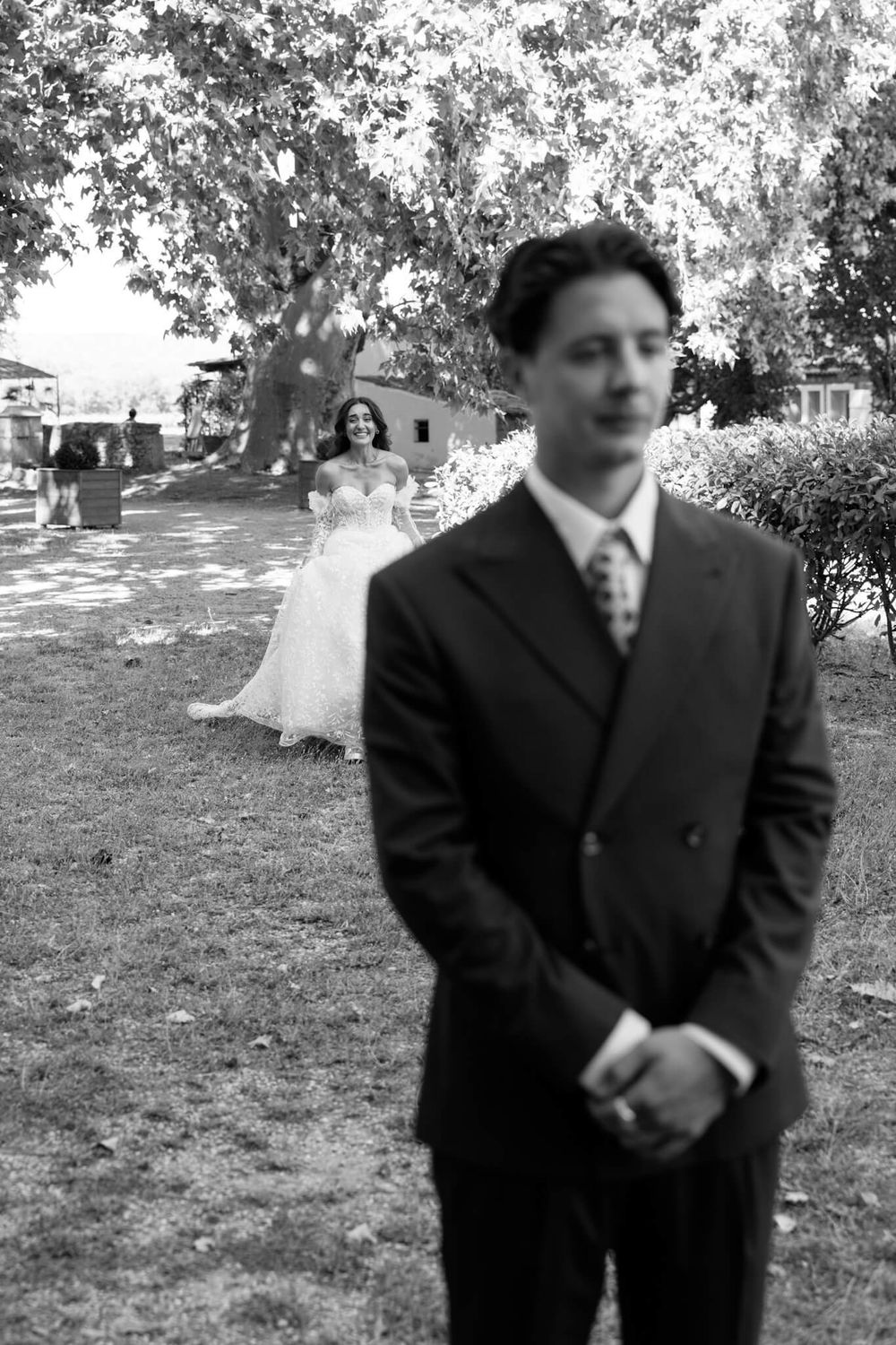 Black and white photo of a groom in a suit standing in foreground as his bride in a wedding dress approaches from behind.