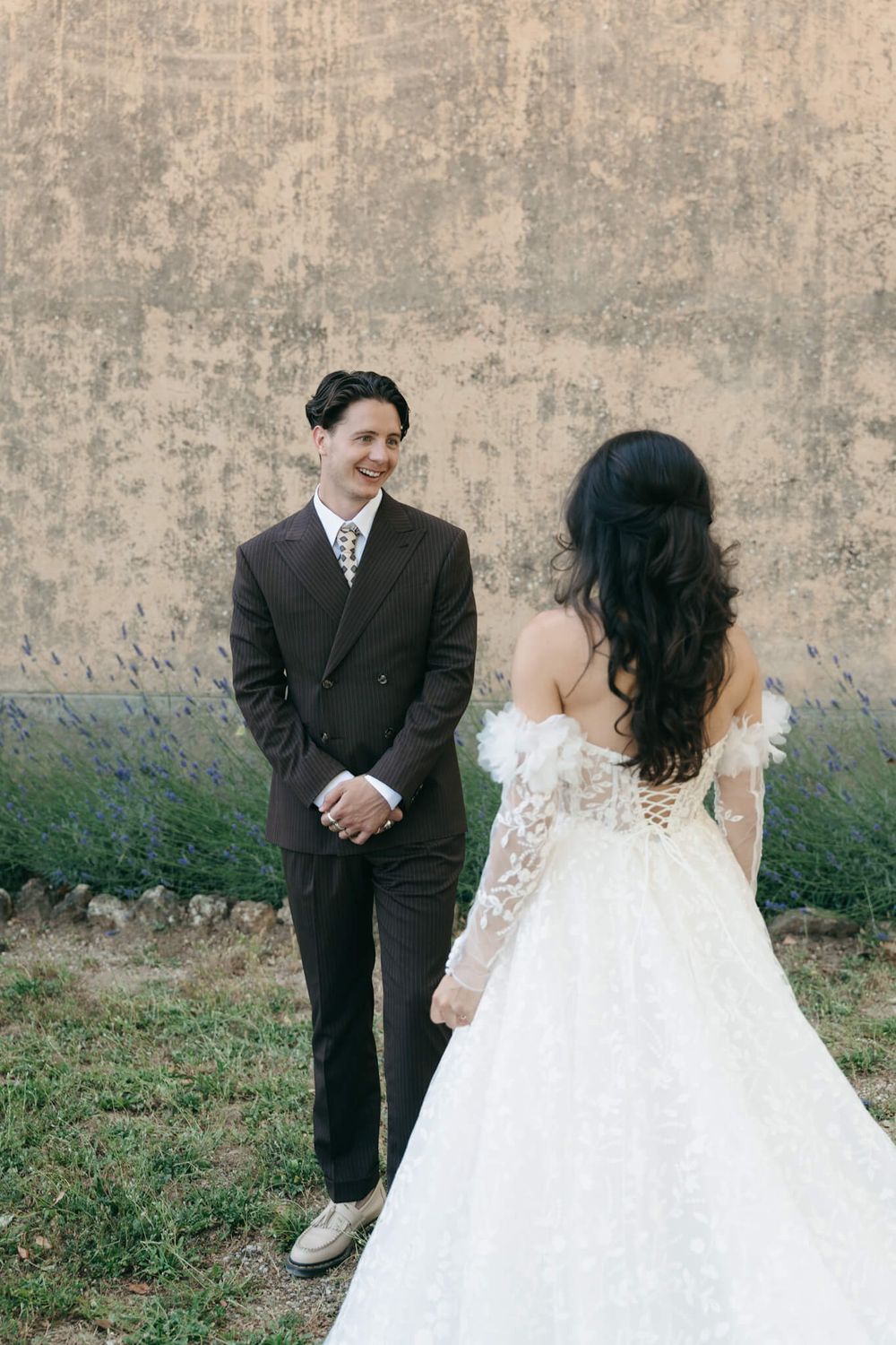 A smiling groom in a dark suit faces his bride in a lace off-shoulder wedding gown outdoors.
