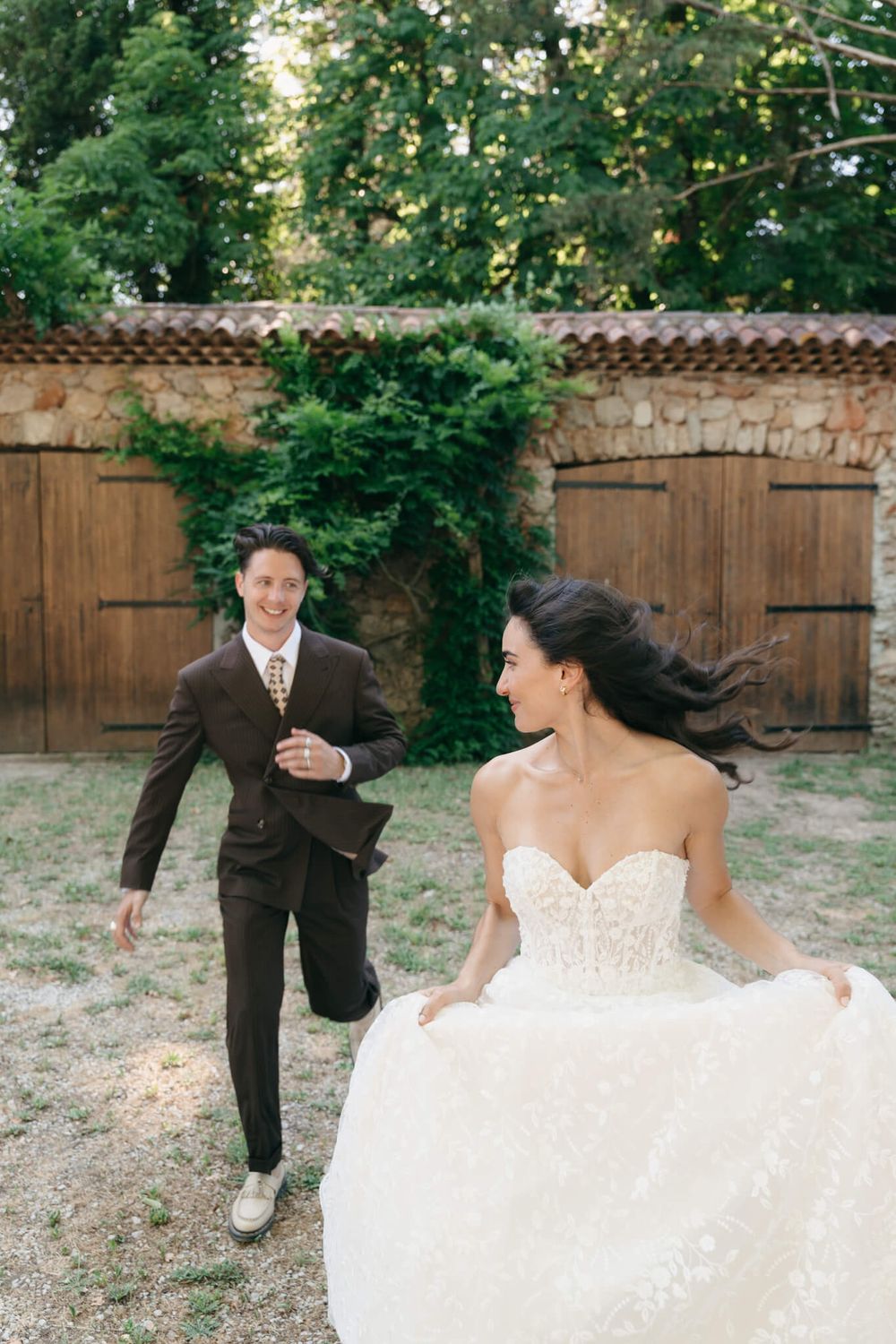 A joyful bride in a white ballgown runs toward her groom in a brown suit outdoors near a rustic stone wall.
