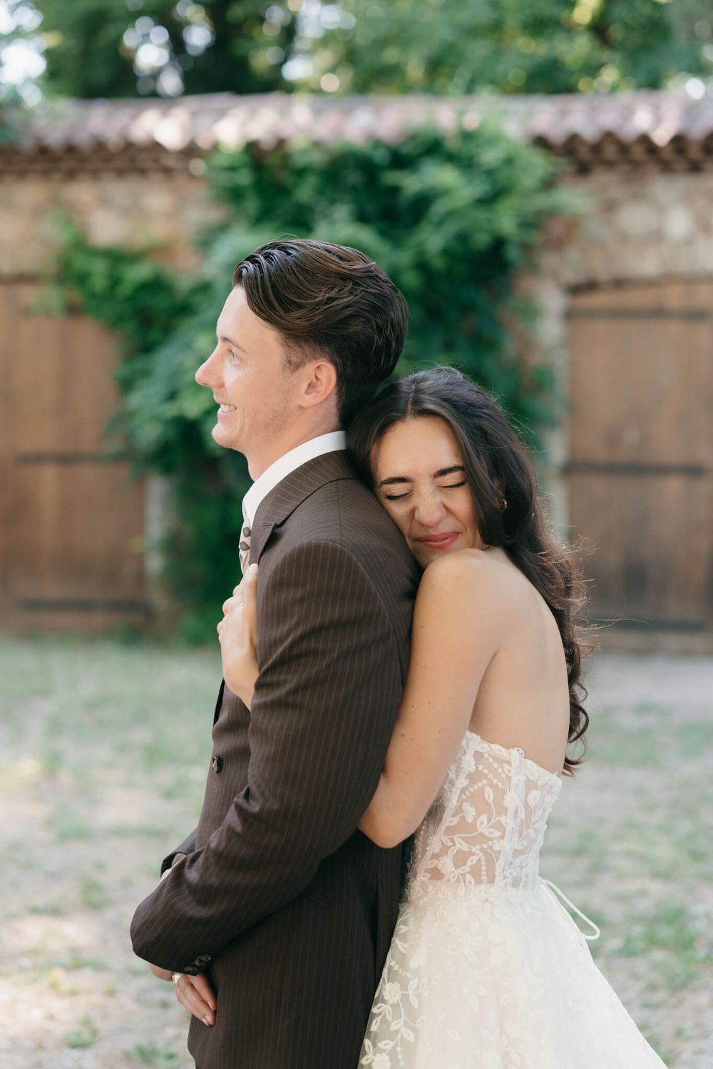 Bride in lace gown embraces groom in brown suit from behind, both smiling outdoors at a garden wedding venue.