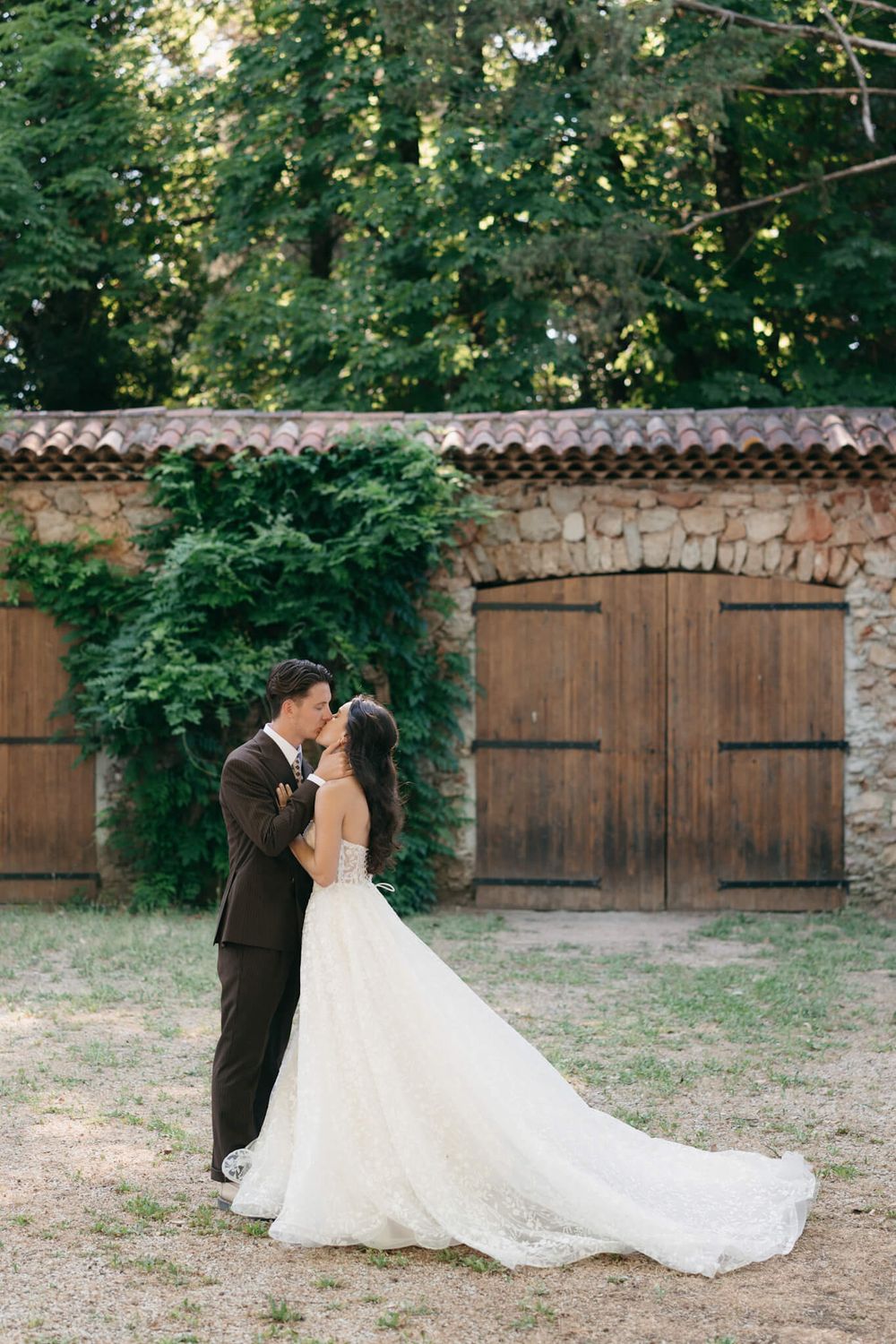 Bride and groom share a romantic kiss outside a rustic stone building with wooden doors and lush greenery.