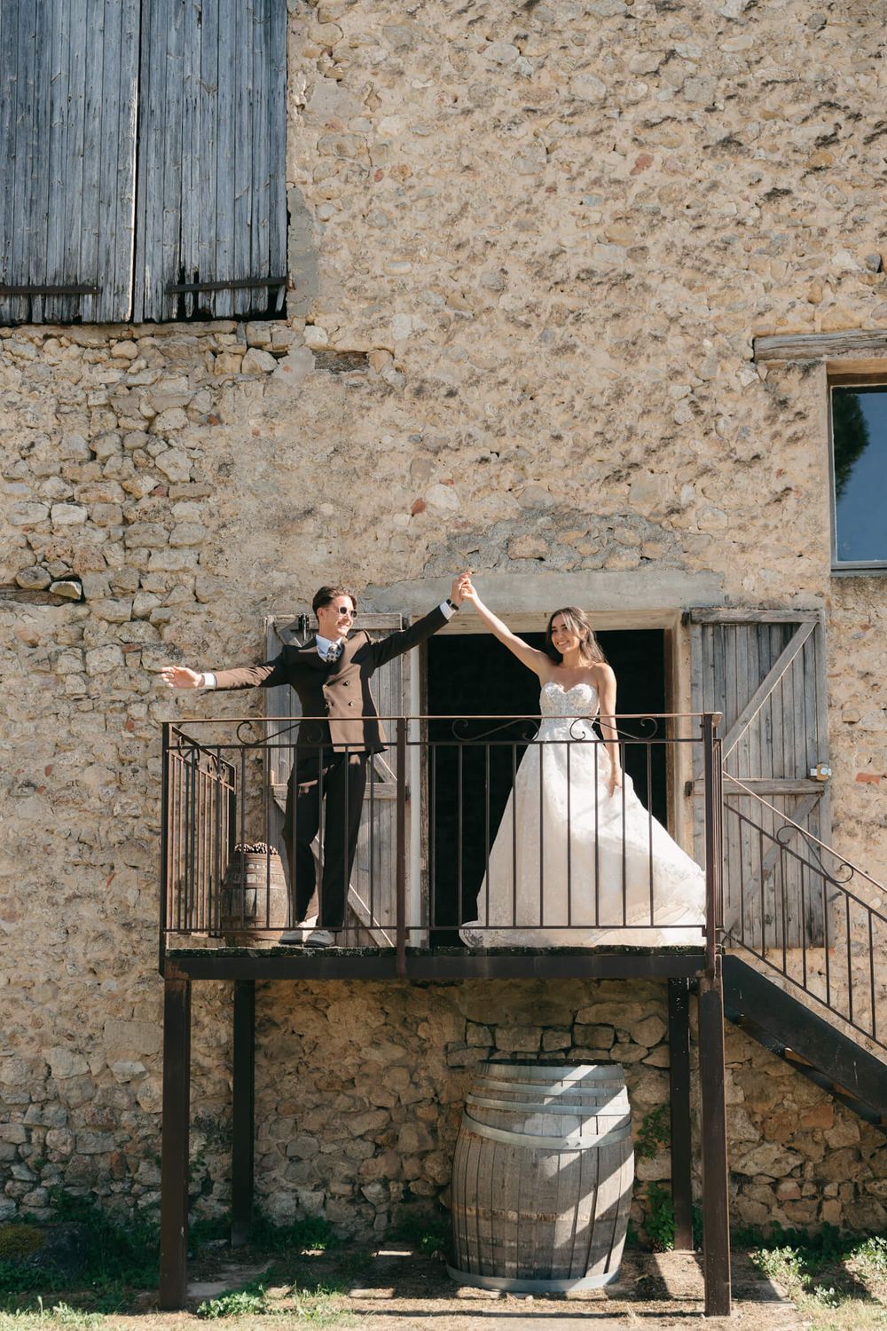 Joyful newlyweds celebrate on a rustic stone building balcony, bride in white gown and groom in suit, arms raised.
