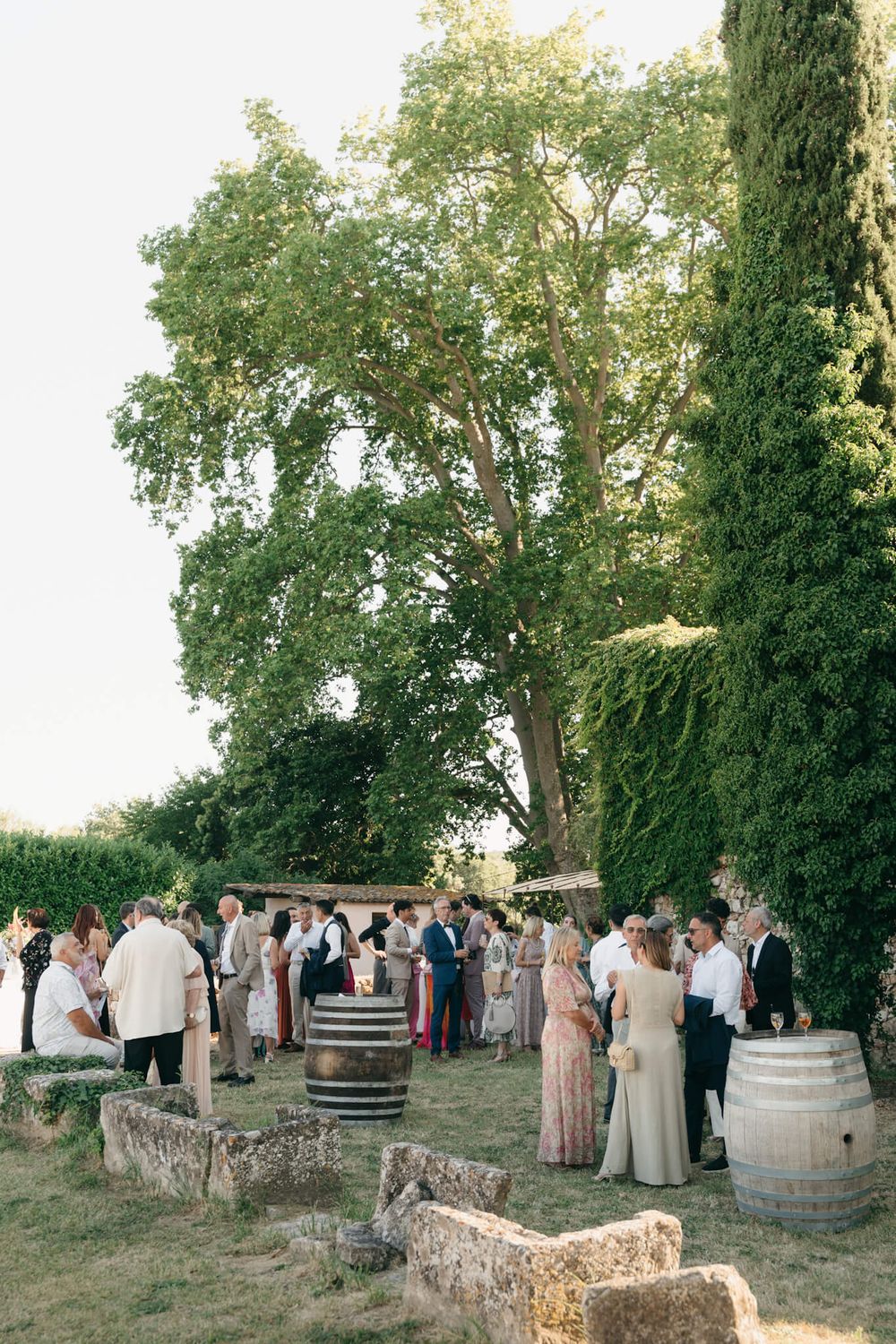 Guests mingle outdoors at a rustic garden wedding reception with wine barrels and lush green trees.
