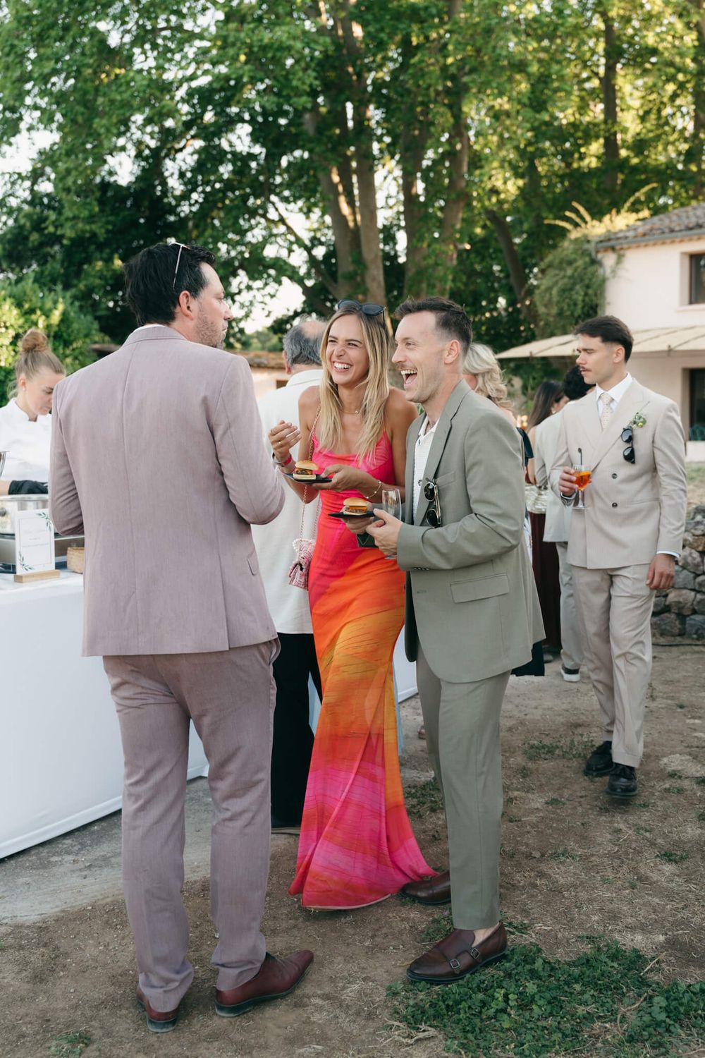 Guests in elegant attire socializing outdoors at a wedding reception, woman in colorful dress laughing with two men in suits.