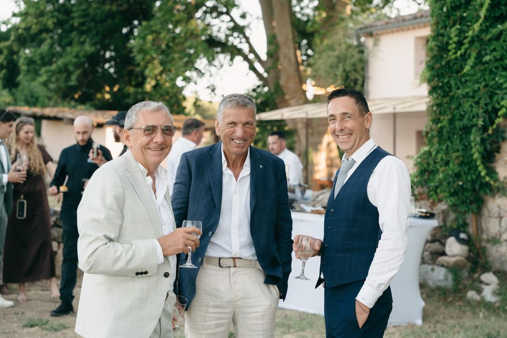 Three men smiling and holding wine glasses at an outdoor wedding reception in a garden setting.
