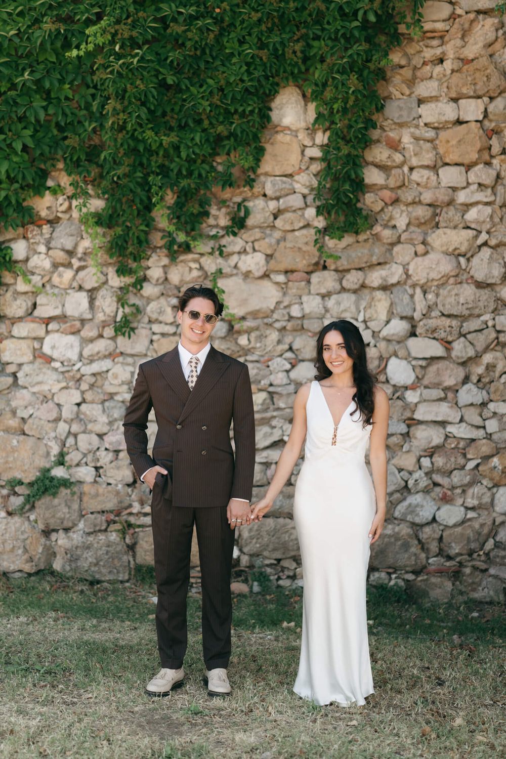 Bride in white gown and groom in brown suit hold hands before a rustic stone wall with ivy.