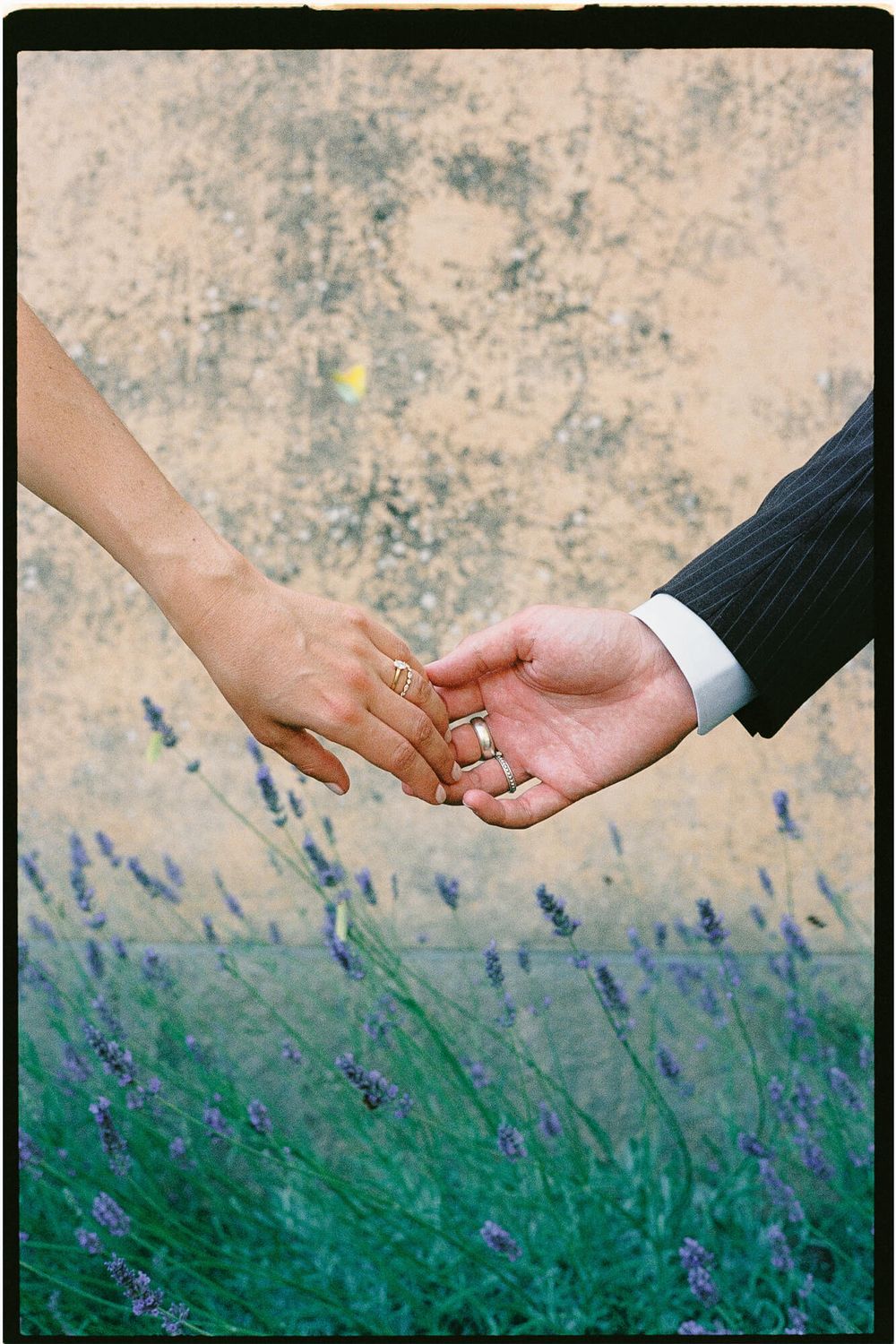 Newlywed couple holding hands showing wedding rings with lavender flowers and rustic wall backdrop.