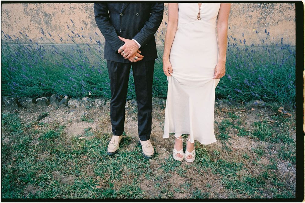 Bride in white dress and groom in black suit holding hands outdoors, showing only their lower bodies and shoes.