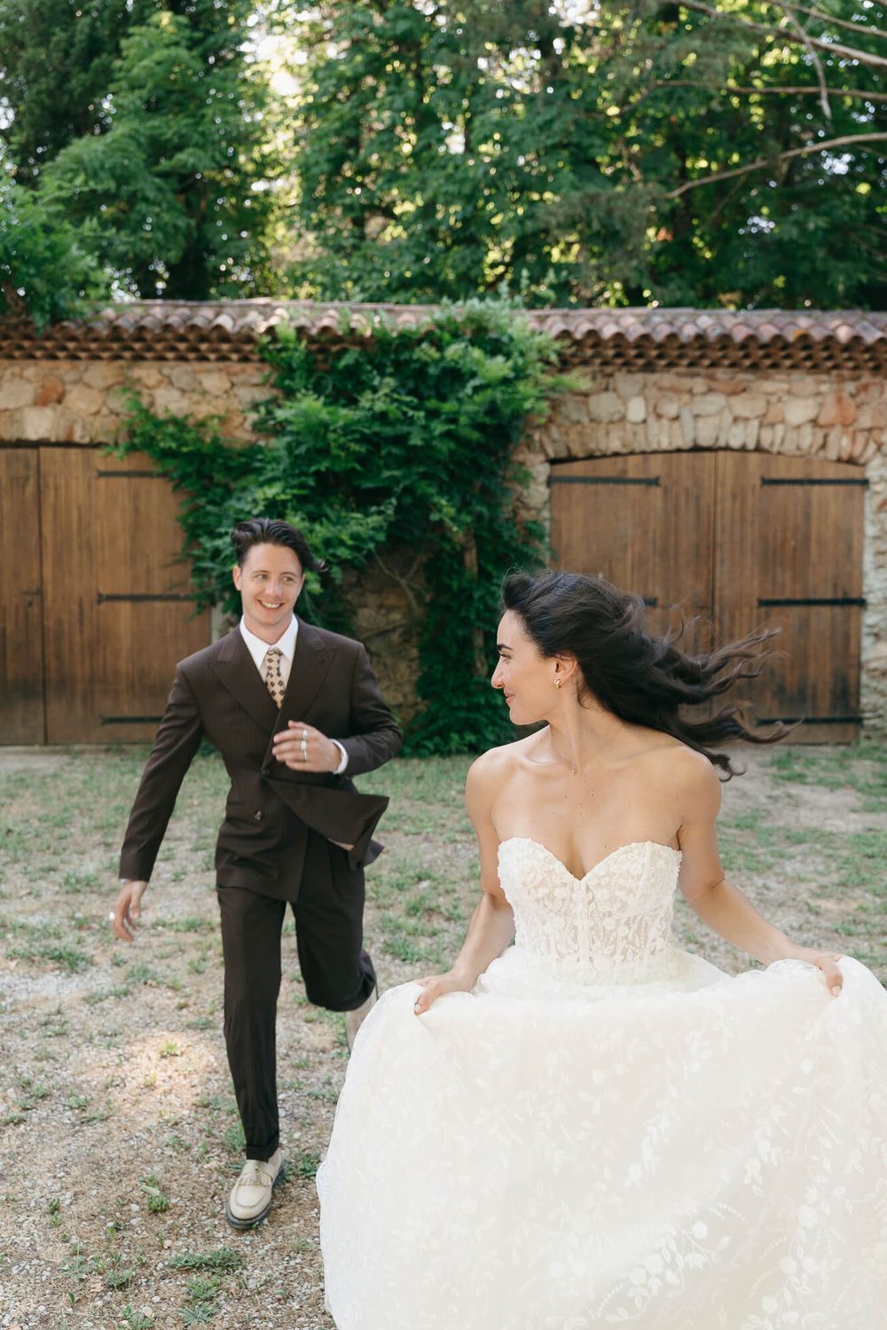 Bride in white gown and groom in brown suit running and laughing together outdoors near rustic stone wall.