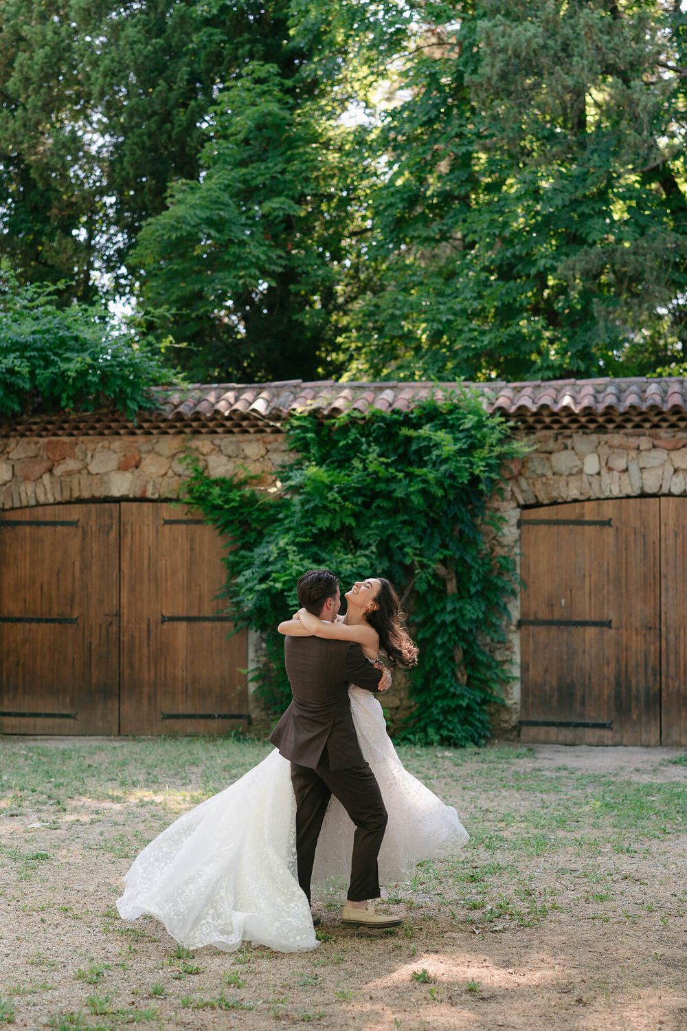 Bride and groom share a romantic dance outdoors near rustic wooden doors surrounded by lush green trees.