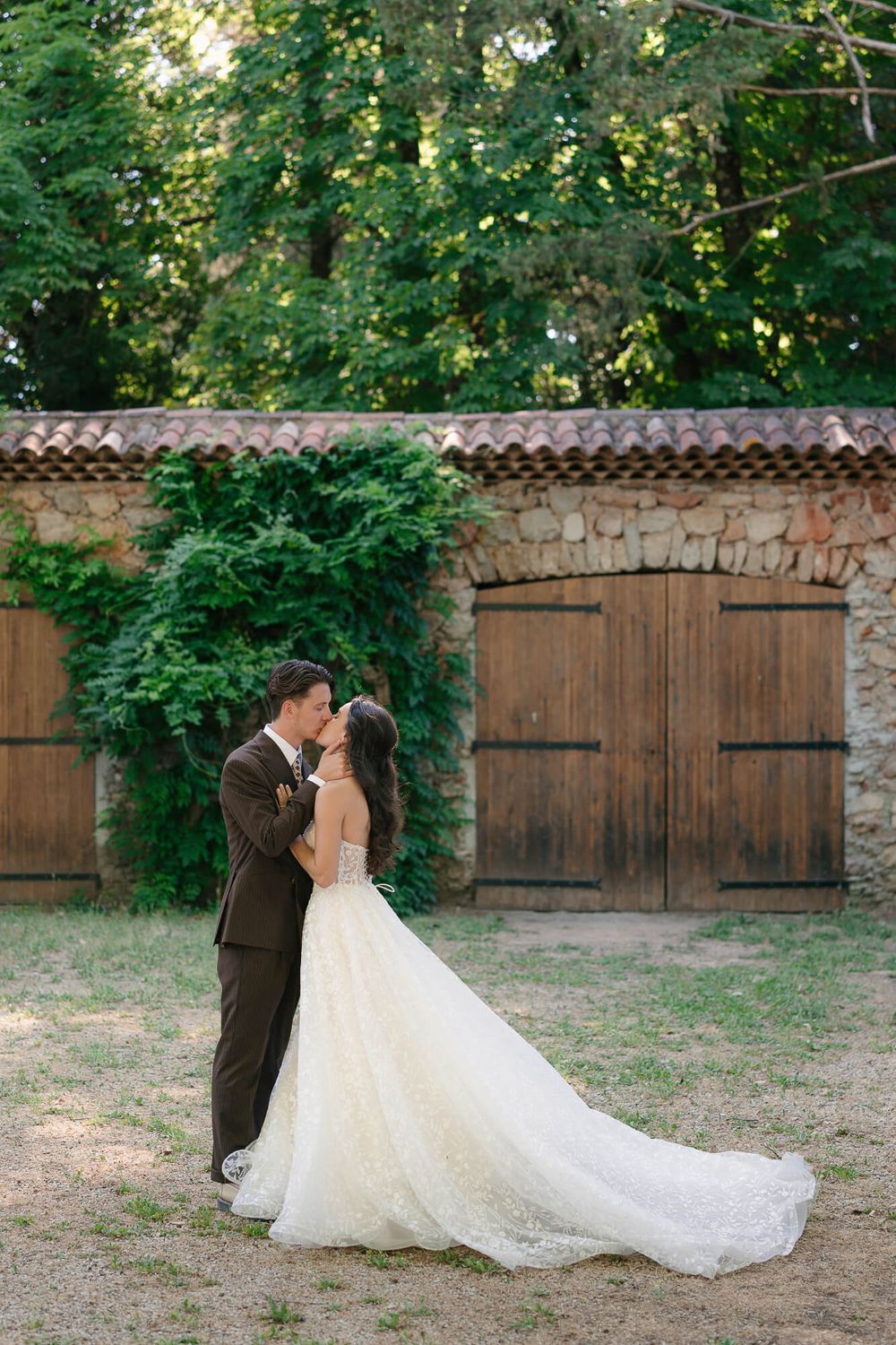 A bride and groom share a romantic kiss in front of a rustic stone building with wooden doors and lush greenery.