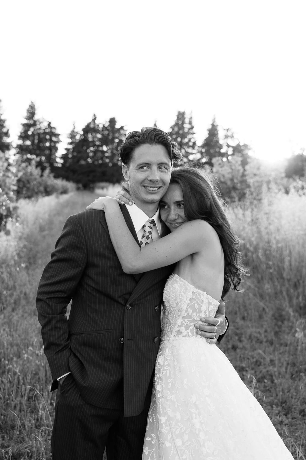 Black and white photo of a bride in lace gown embracing a groom in pinstripe suit outdoors.