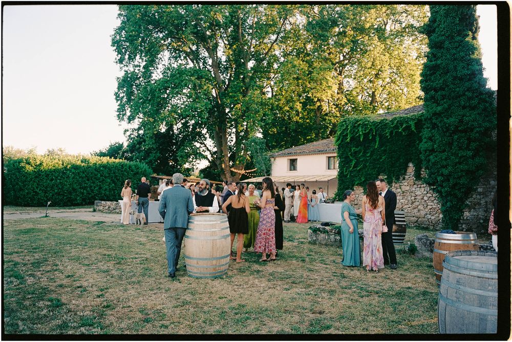 Guests mingle outdoors at a rustic wedding reception with wine barrels and trees in golden evening light.