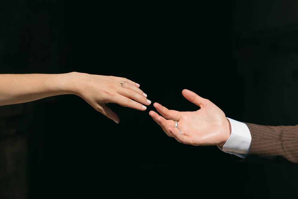 Two hands reaching toward each other against dark background, woman wearing engagement ring, man in suit sleeve.